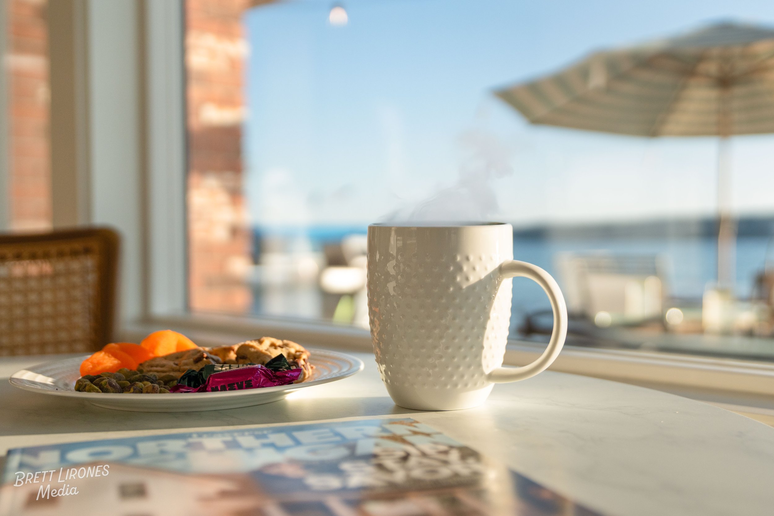 A white mug with steam rising from it, placed on a table near a window. Behind the mug, there is a plate with assorted breakfast items including fruit, cookies, and wrapped candies. Outside the window, there is a clear blue sky and a large umbrella.