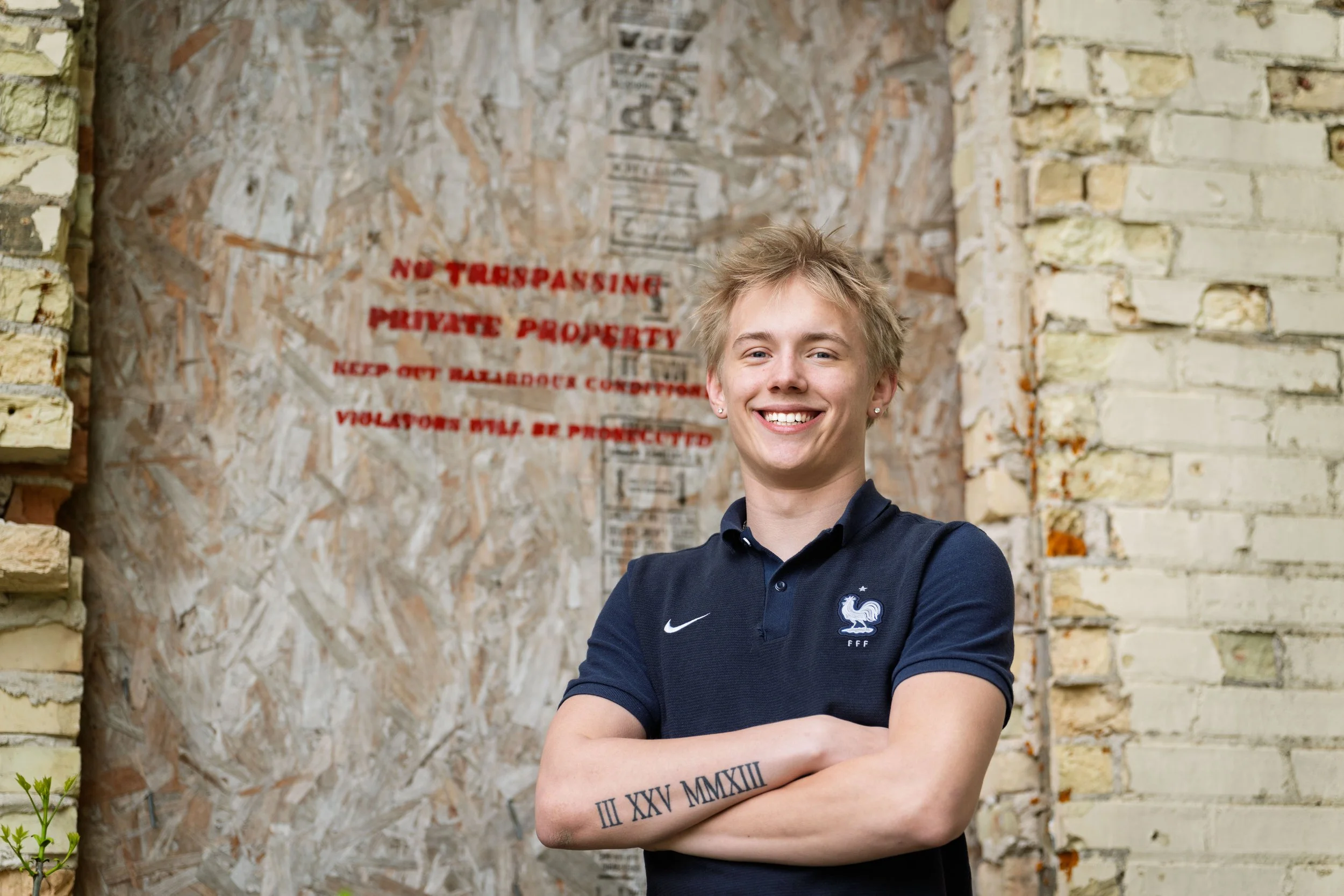 A young woman with short blonde hair and earrings, wearing a black polo shirt with a French football emblem, smiling with arms crossed, standing outdoors near a rustic brick wall with a faded sign that says 'No Trespassing Private Property' in red le