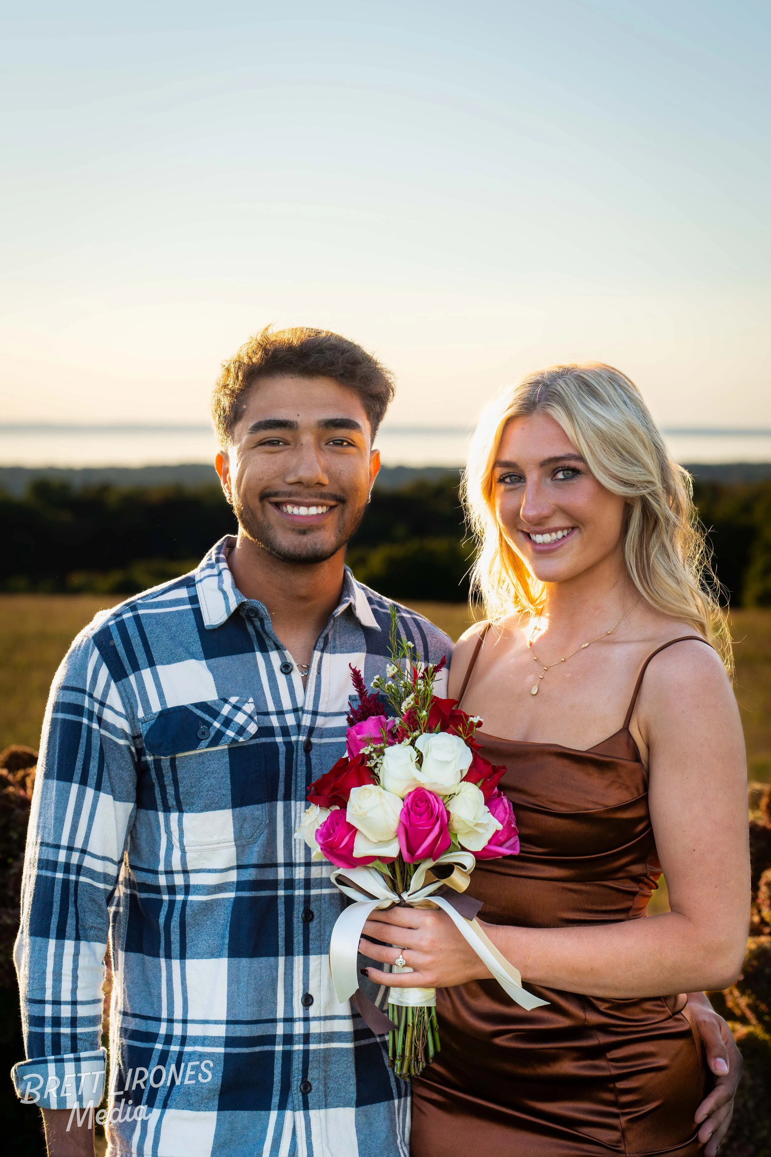 A young man and woman standing outside at sunset, smiling, with the woman holding a bouquet of pink and white roses.
