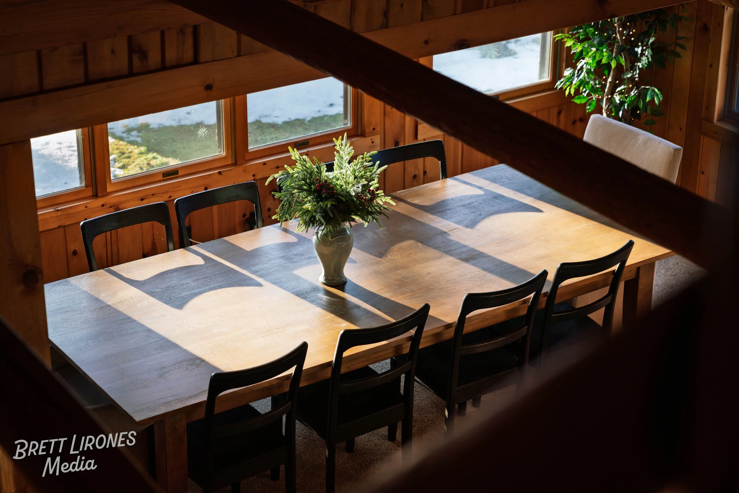 A wooden dining table with six black chairs and a vase of flowers in the center, illuminated by sunlight through windows, inside a cozy wooden room.