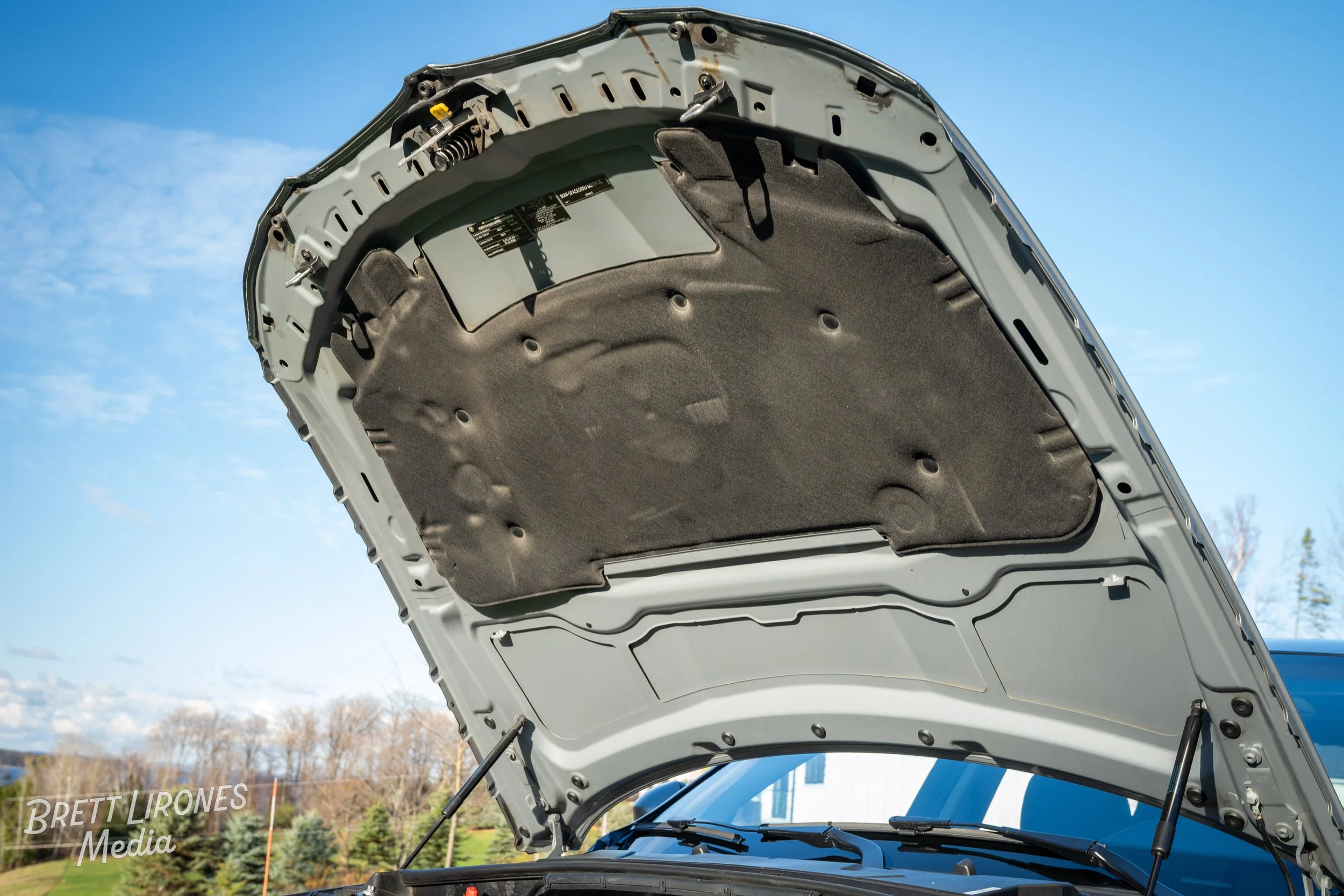 An open car hood showing the underside with insulation padding, hydraulic lift supports, and part of the windshield wipers visible. The background features a clear blue sky and trees.
