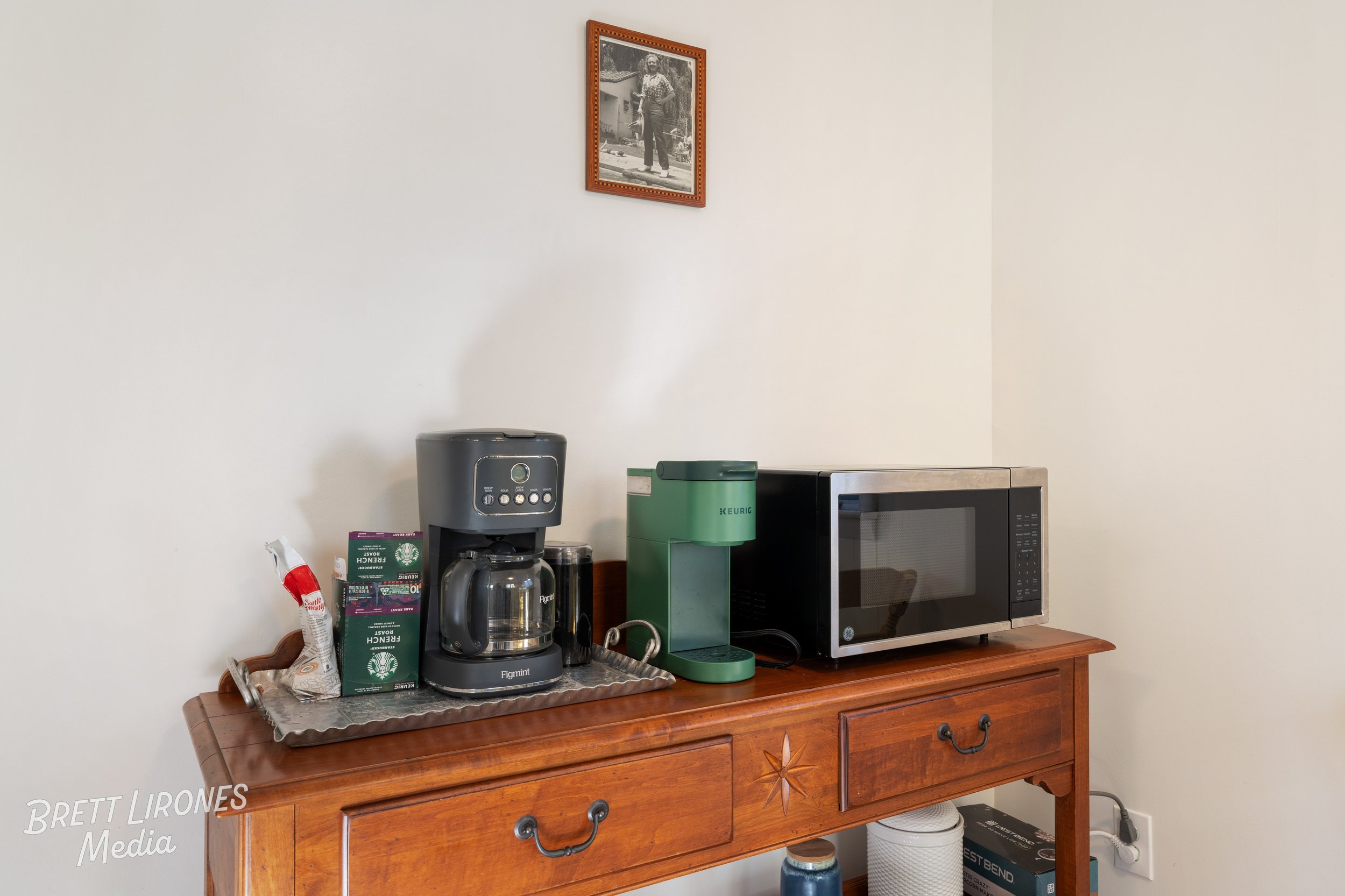 Wooden sideboard on a light-colored wall with a coffee machine, a green Keurig, and a microwave on top. A framed black-and-white photo hangs on the wall above.
