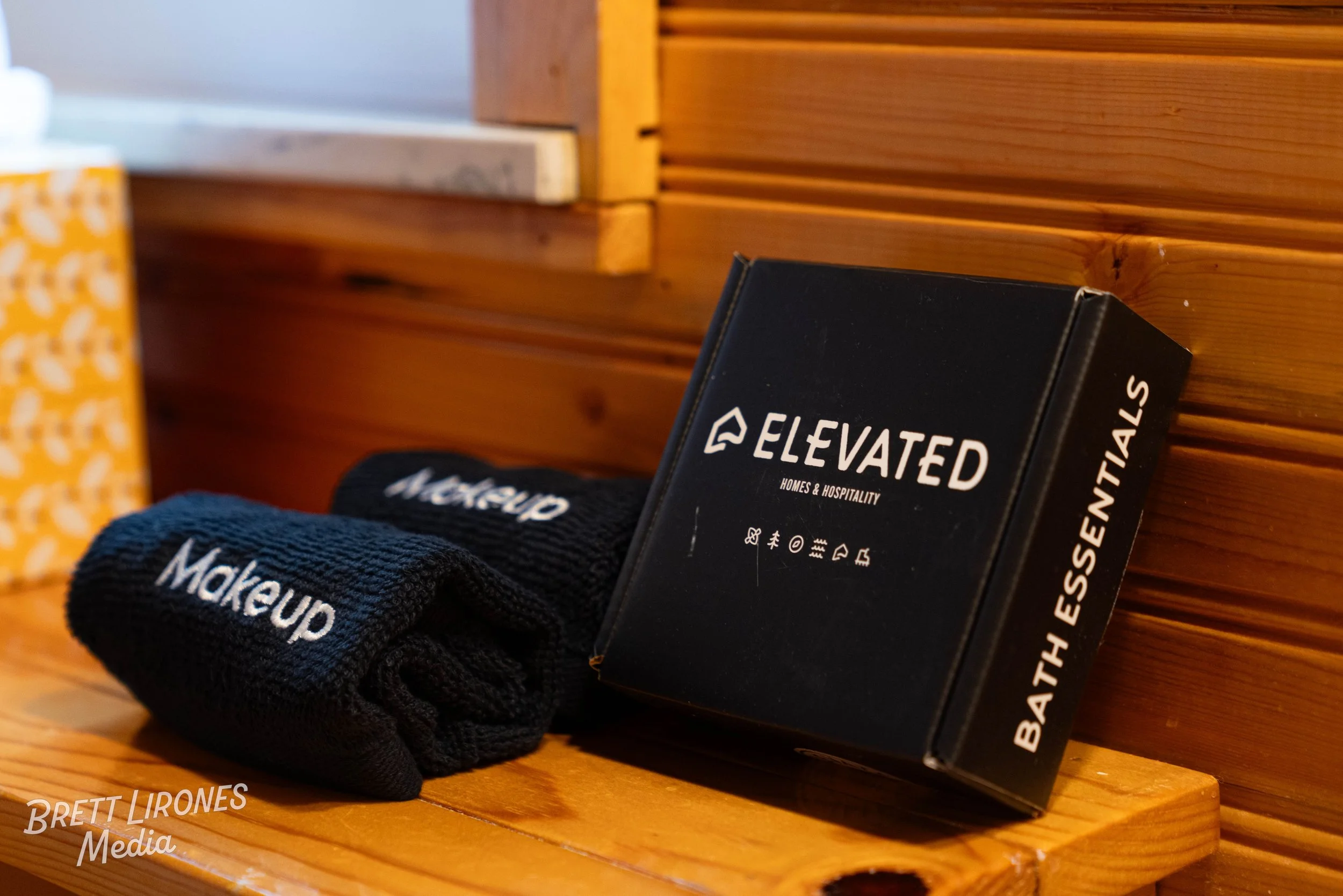 A black box labeled 'Elevated Homes & Hospitality' and a pair of black socks with 'Makeup' written on them are placed on a wooden surface against a wooden wall.