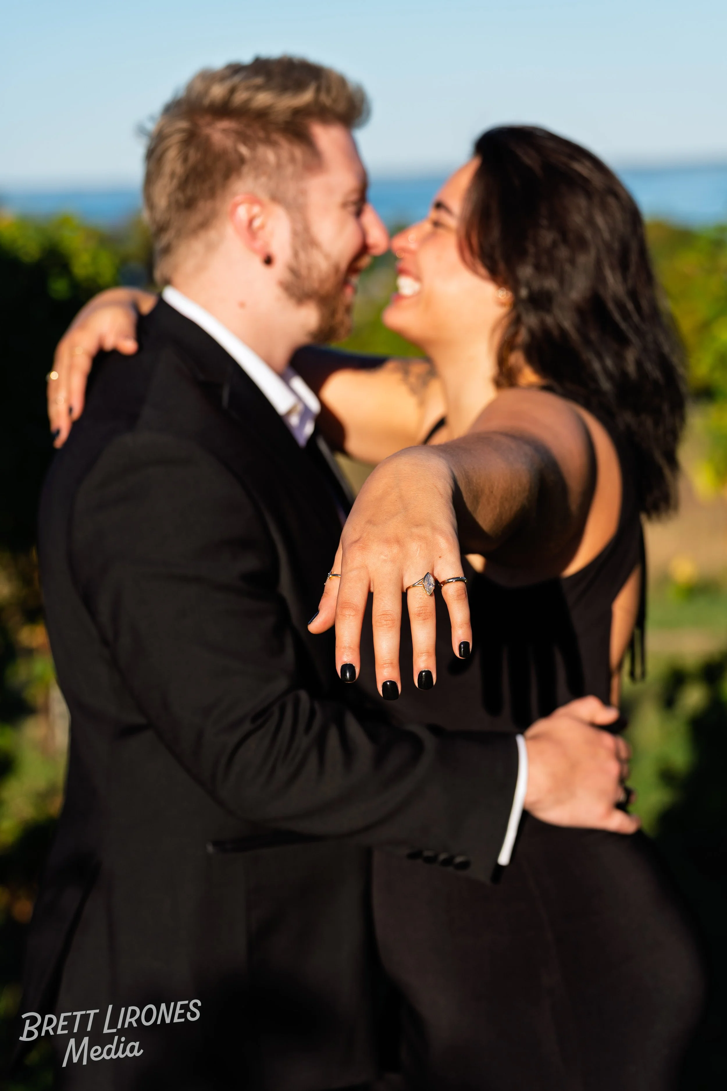 A happy couple embracing outdoors, smiling and looking into each other's eyes, with the woman's hand showing rings and black nail polish.