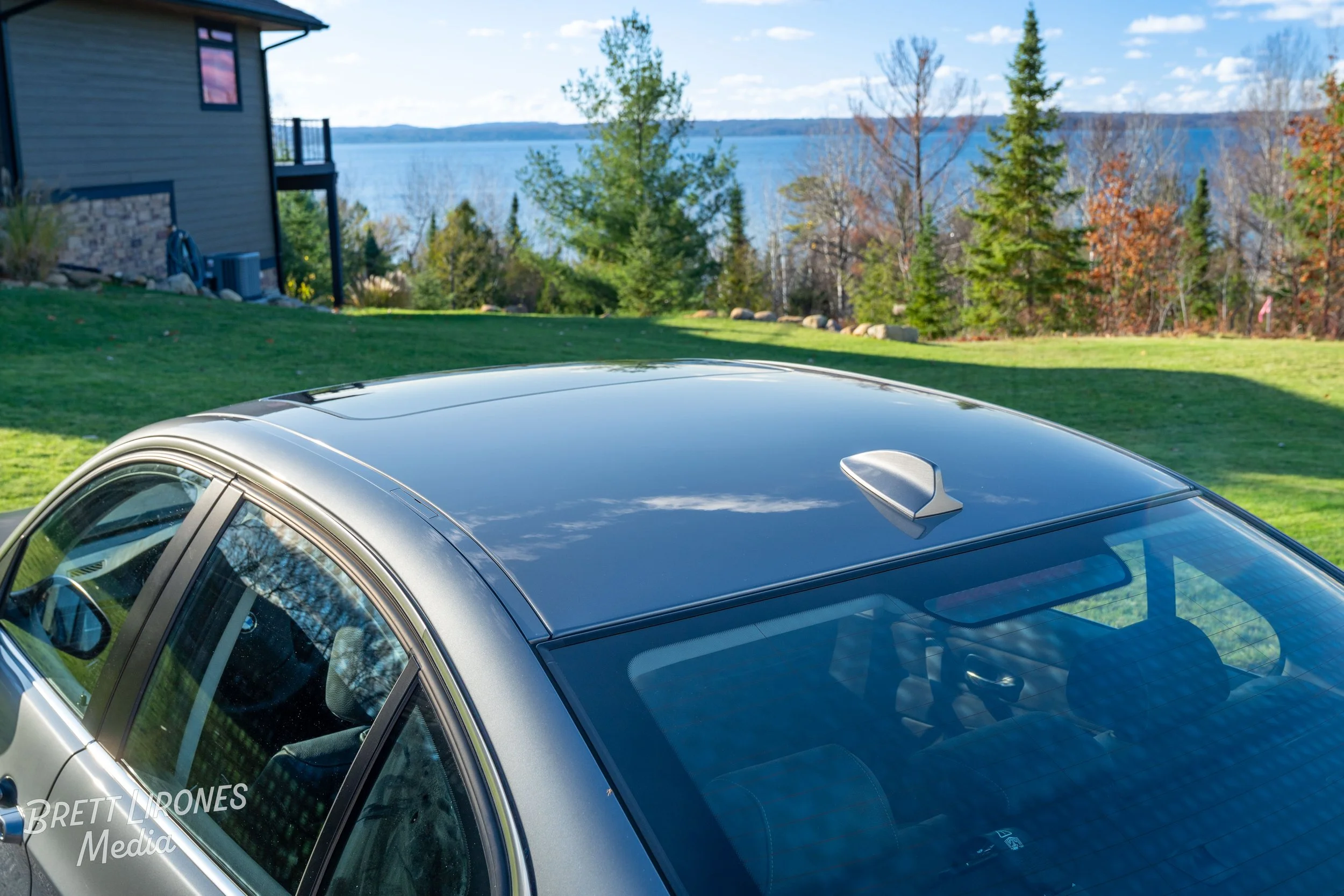 Silver car parked on a grassy area with trees and a lake in the background, blue sky with clouds.