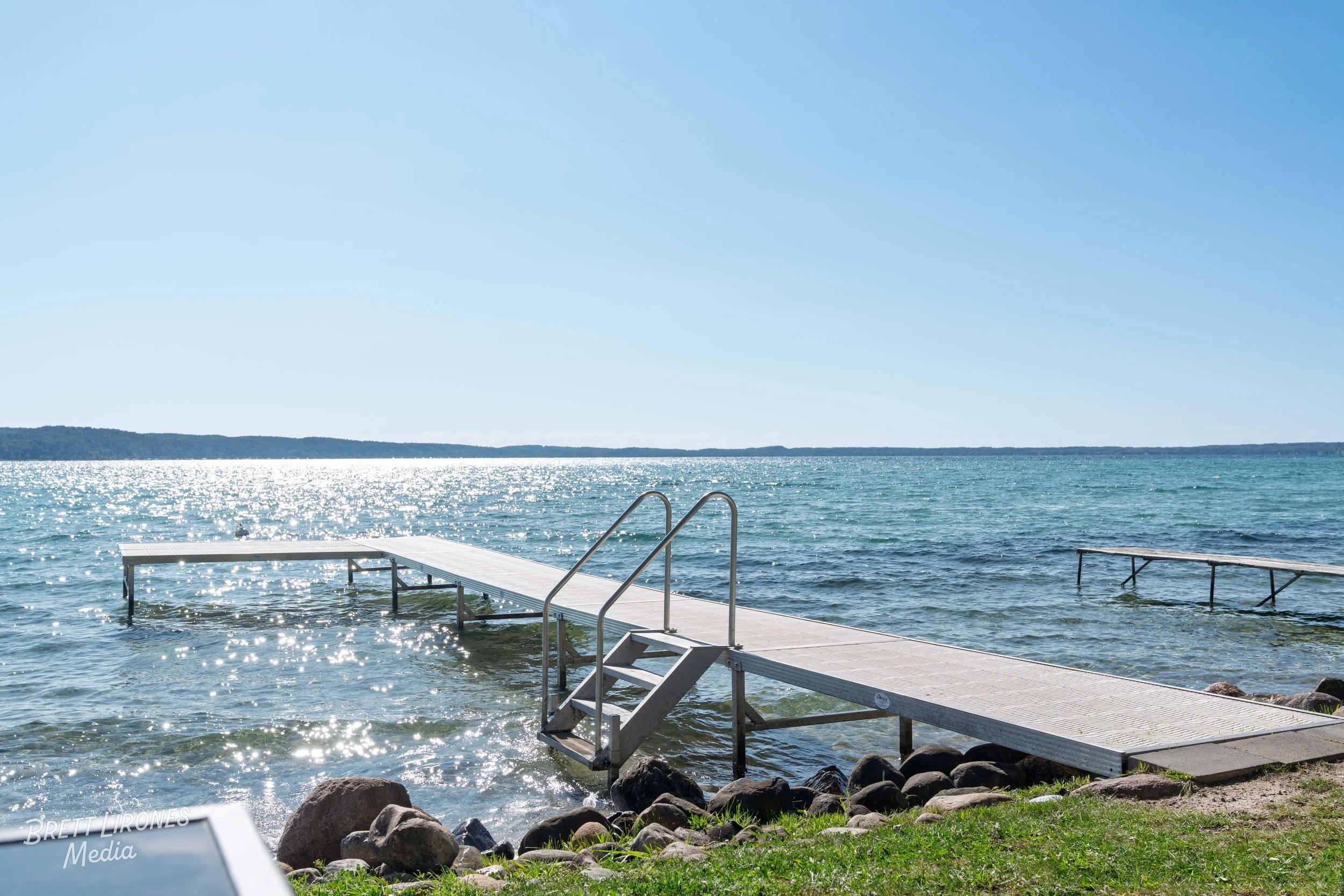 A wooden dock with stairs leading into a calm, sparkling lake, with a clear blue sky overhead and distant shoreline in the background.