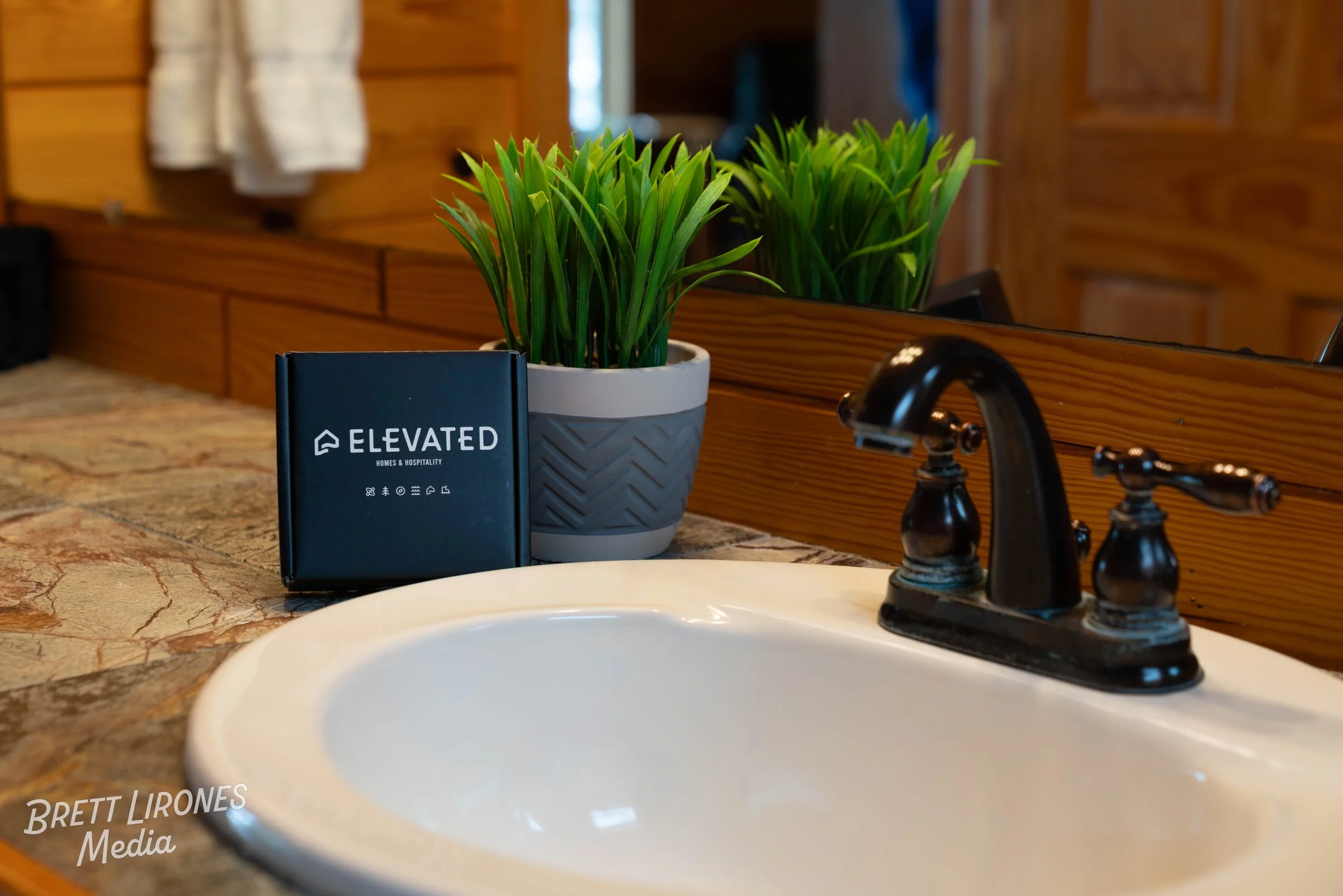A bathroom countertop with a white sink, black faucet, green potted plant, and a black box with the word ELEVATED on it. The background features a mirror, wooden wall, and a towel.