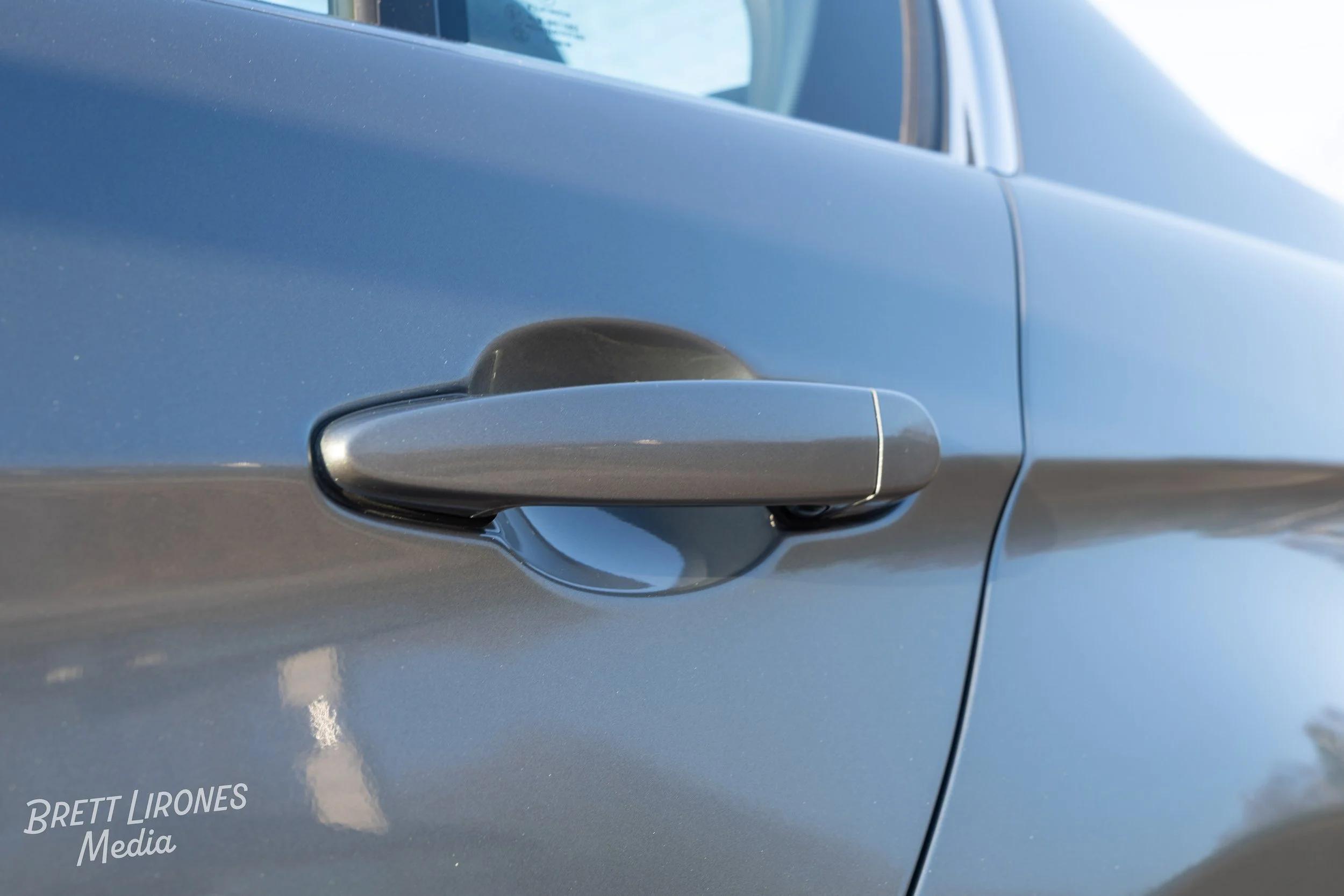 Close-up of a gray car door handle on a blue vehicle with a reflection of the sky and trees.