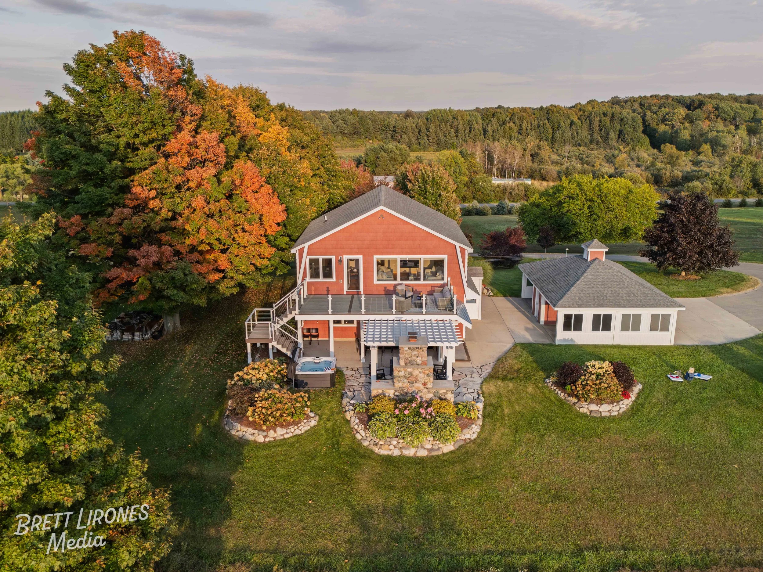 An aerial view of a house with an outdoor patio and hot tub, surrounded by trees with autumn foliage, a garden, and a lawn, in a rural landscape.