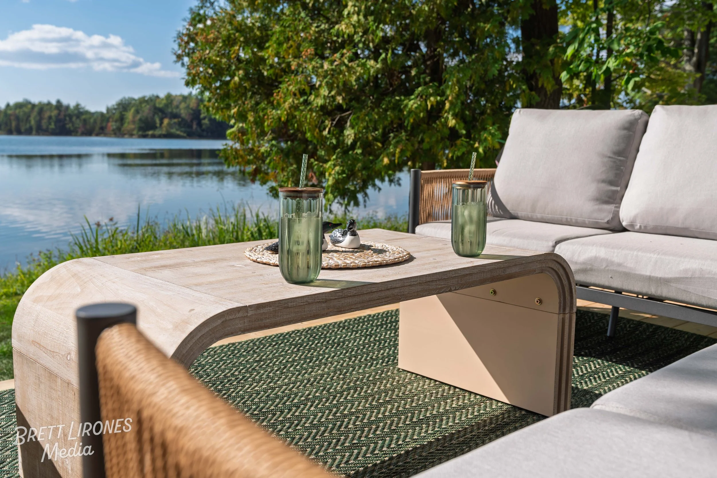 Outdoor patio furniture by a lake with a wooden table, glass of water, and decorative ducks, surrounded by trees and grass on a sunny day.