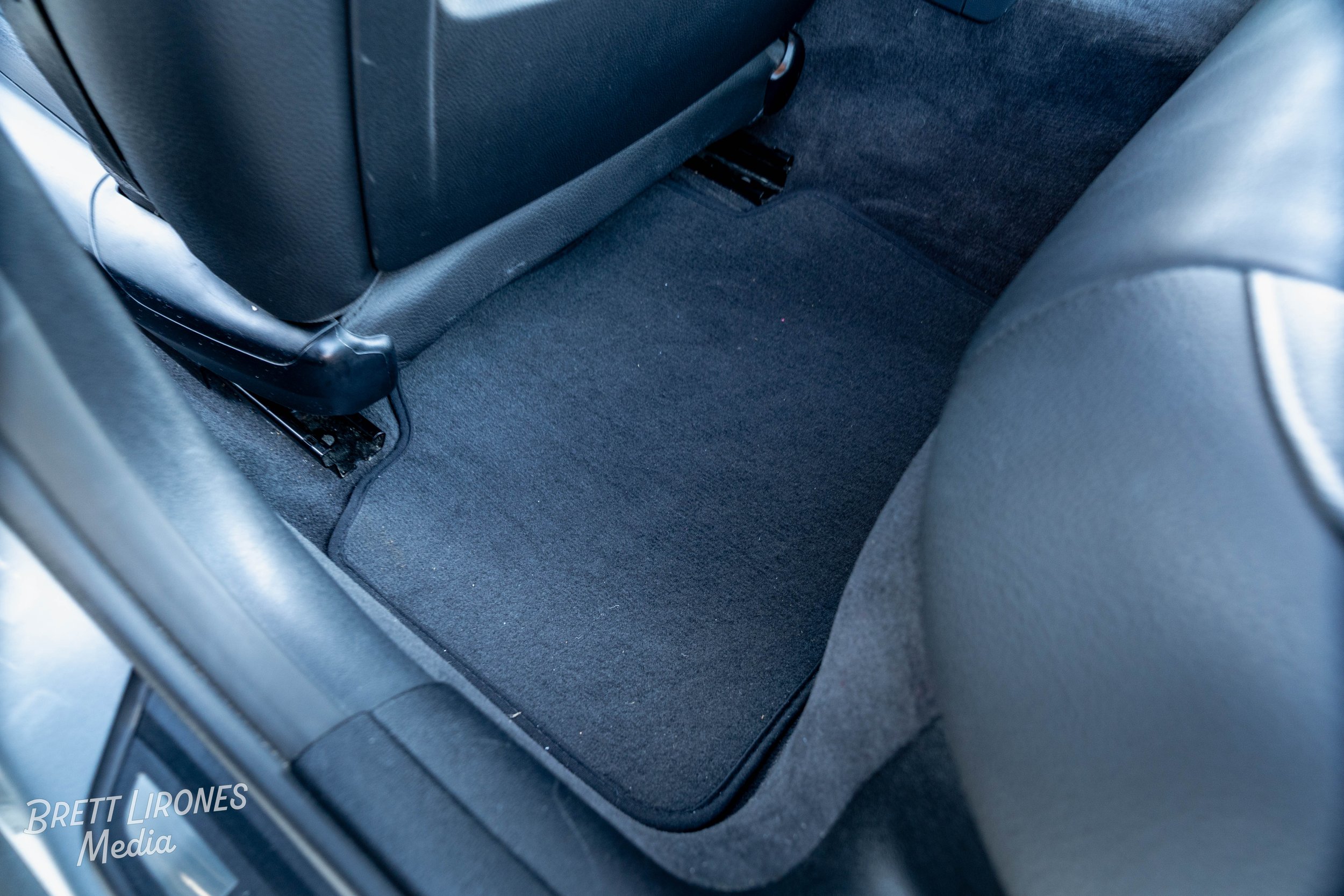 The back seat floor area of a car with a black carpeted mat and a black seatback.