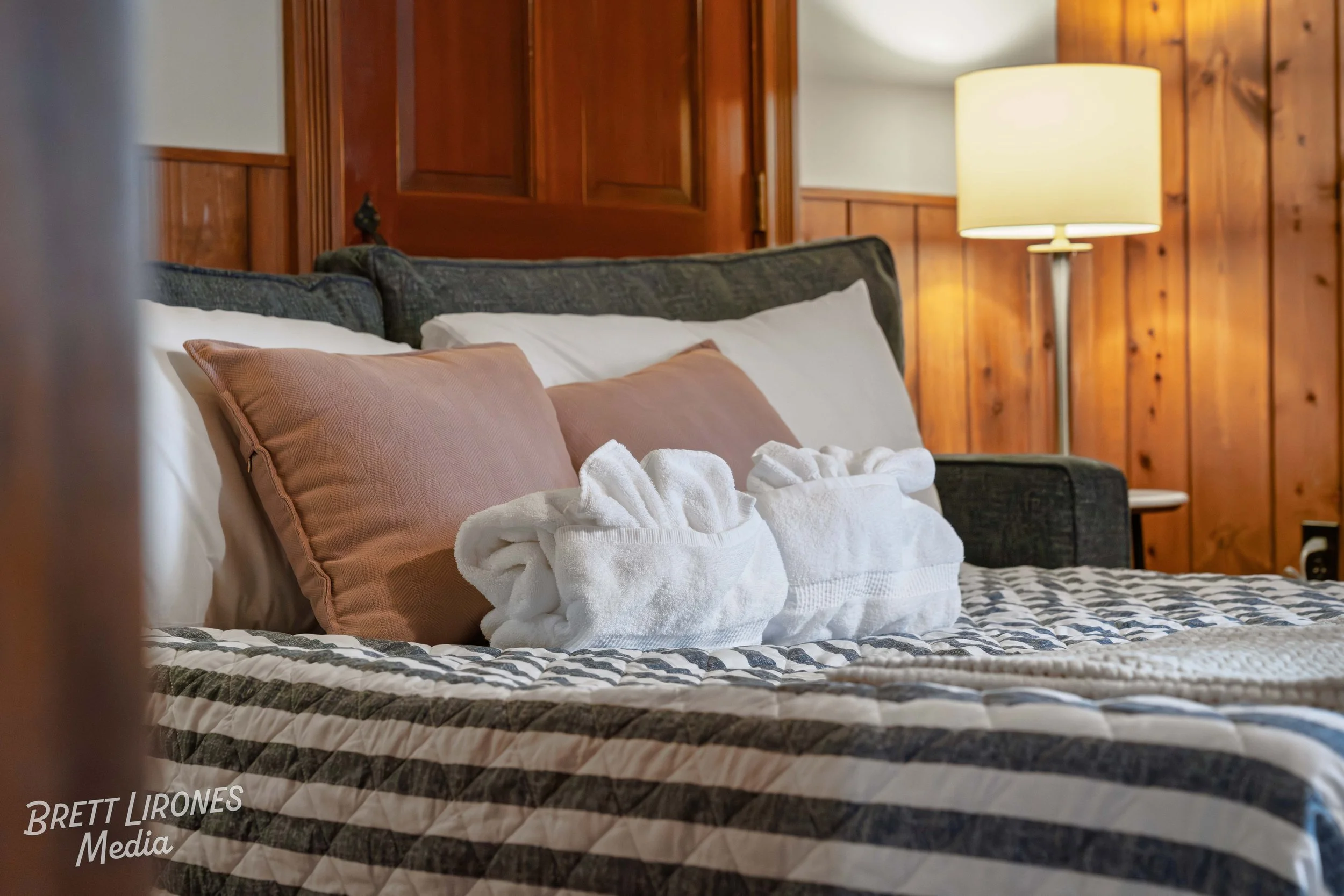 A neatly made bed with striped bedspread, pink pillows, and two towel sculptures shaped like swans, in a room with wooden paneled walls and a standing lamp.