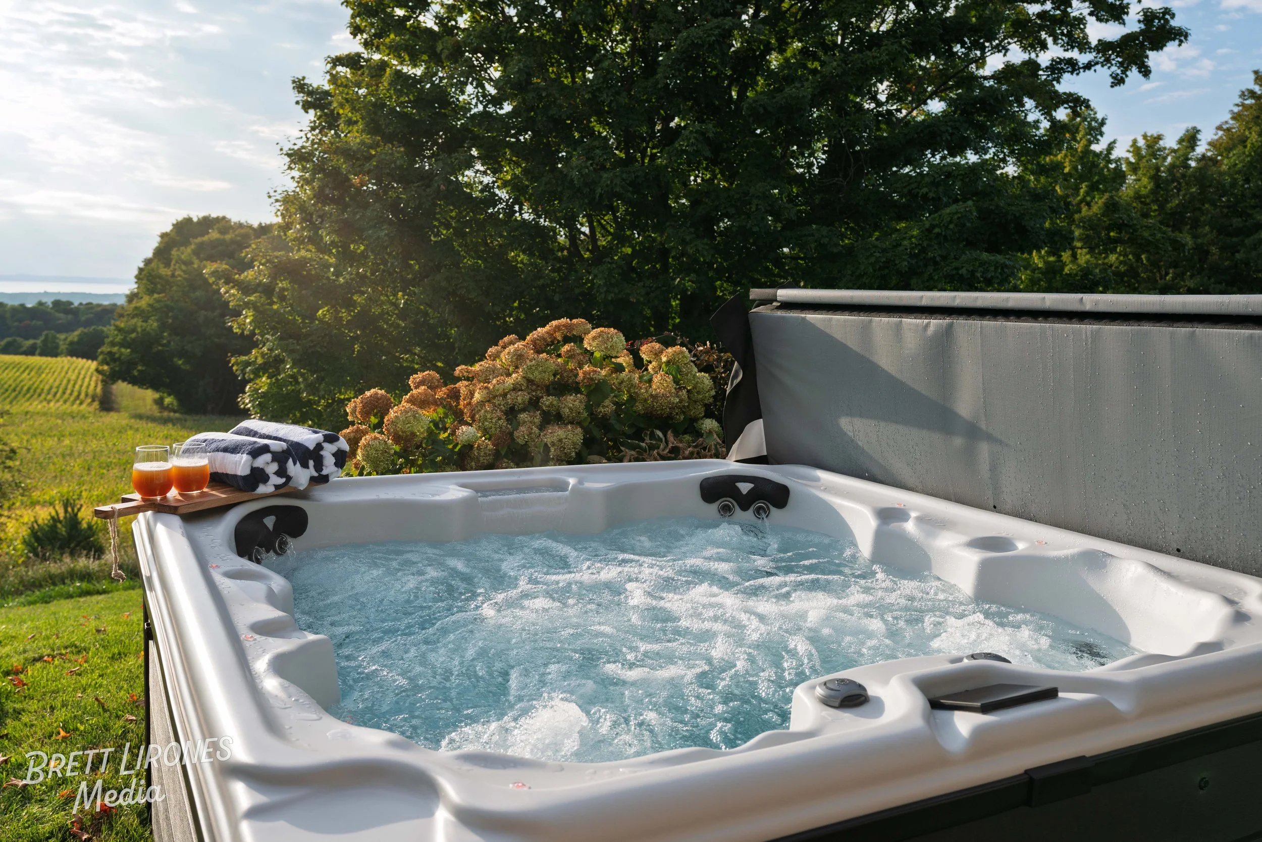 A hot tub filled with bubbling water outdoors on a grassy area, with a tray holding two glasses of drinks and folded towels, surrounded by trees and a scenic landscape.