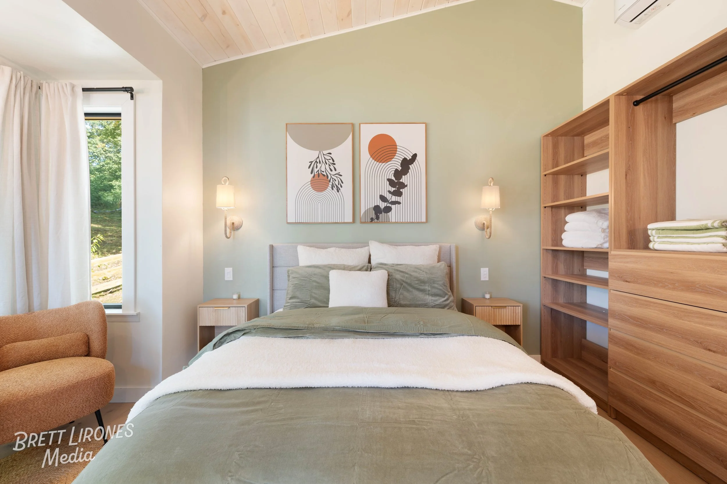 Bedroom with bed, wooden nightstands, abstract artwork, wall-mounted lamps, open wooden shelving with folded towels, beige armchair, window with white curtains, green wall, and wood-paneled ceiling.