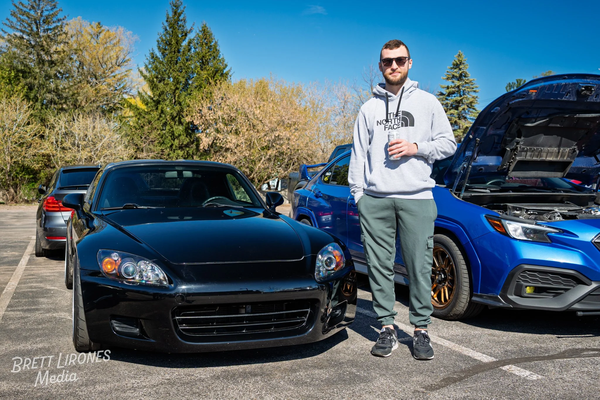 A man with sunglasses wearing a grey hoodie and green joggers standing in a parking lot next to a black sports car and a blue Subaru with its hood open, with trees and a blue sky in the background.