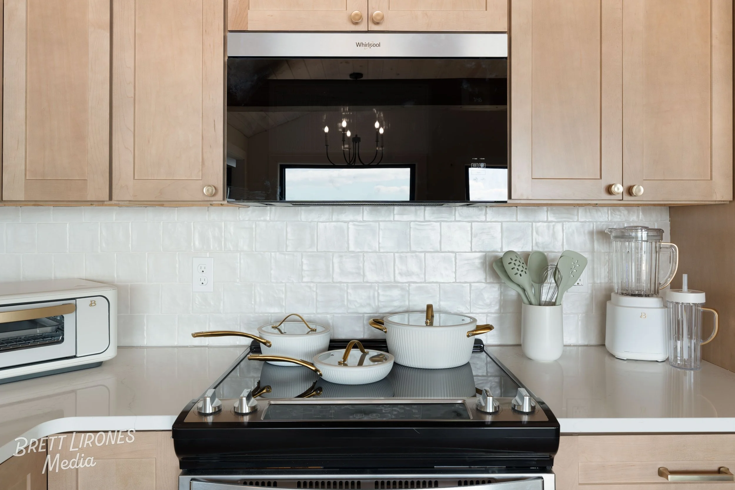 Kitchen countertop with white pots and utensil holder, blender, and toaster, wooden cabinets, microwave, and a black stove with brass handles.