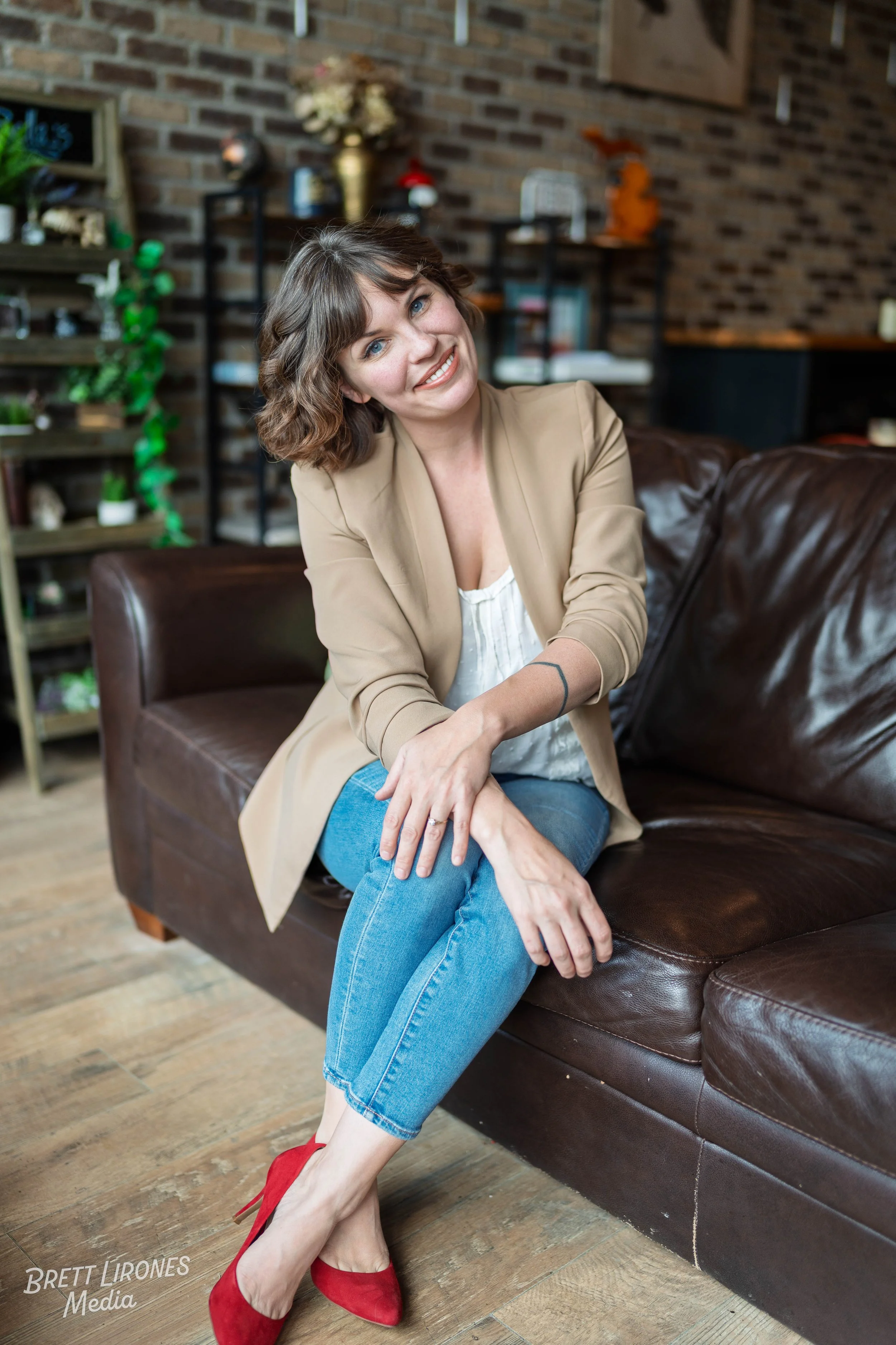 A woman with brown, wavy shoulder-length hair and blue eyes sitting on a dark brown leather couch in a cozy room with a brick wall background. She is smiling and wearing a beige blazer, white blouse, blue jeans, and red high heels.