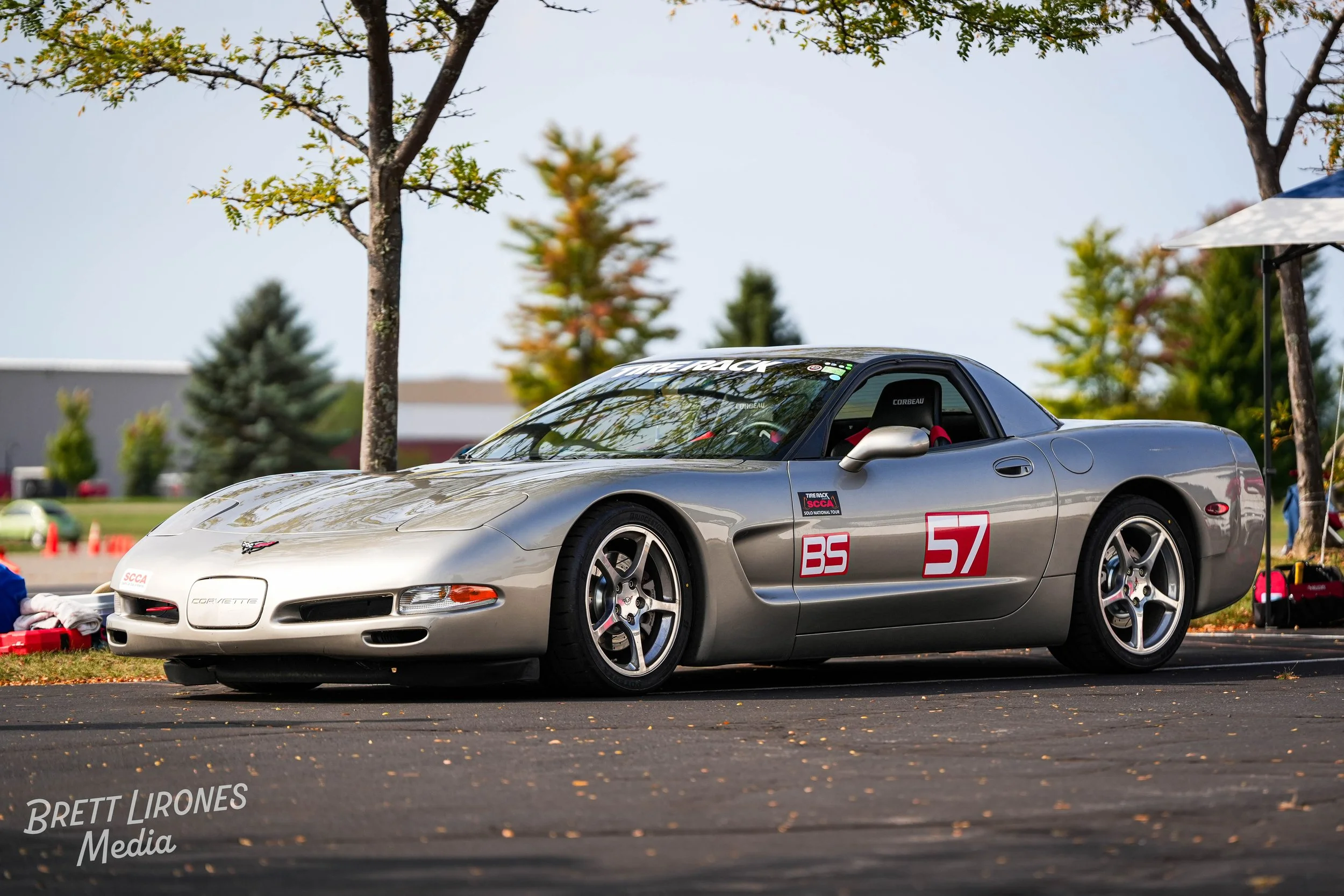 A silver Corvette race car with the number 57 is parked on the track during a race event, surrounded by trees and equipment.