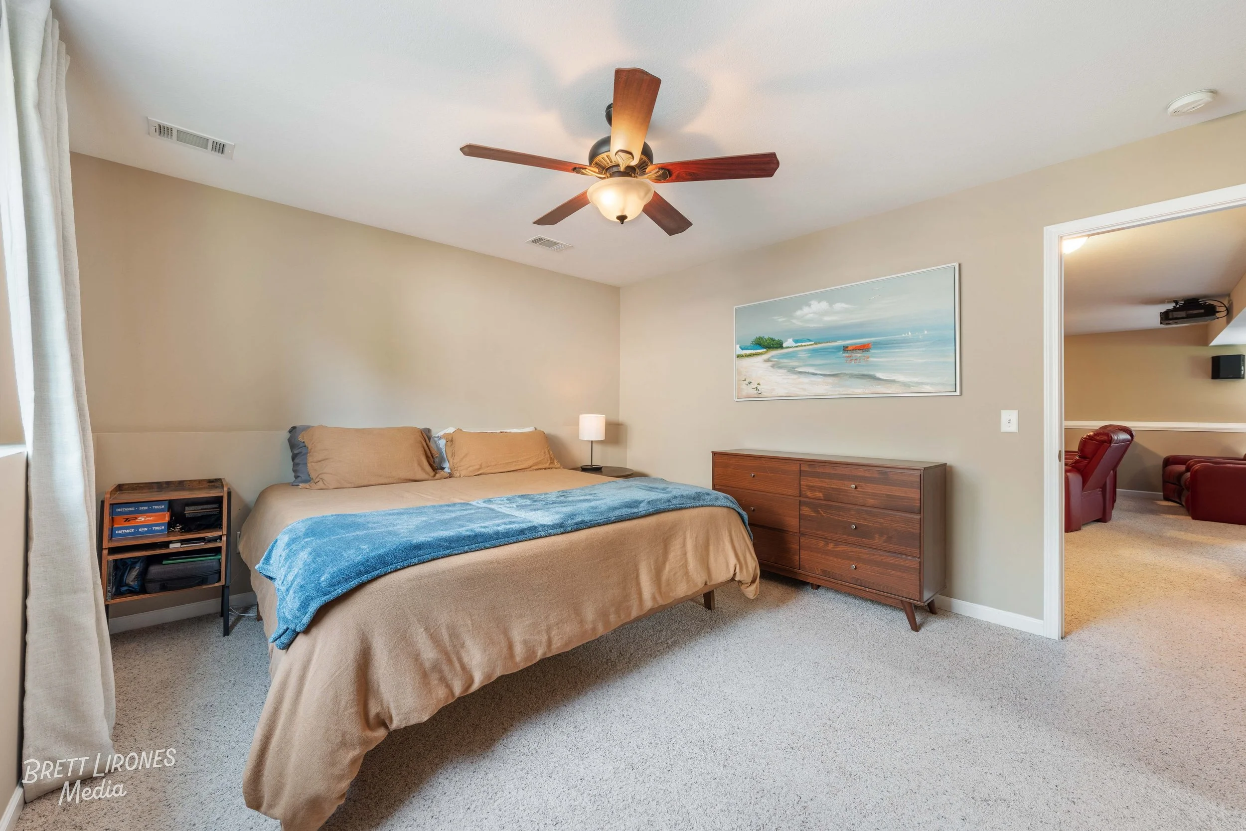 A bedroom with beige walls and carpet, featuring a bed with beige and blue bedding, a small nightstand with books, a wooden dresser, and a large wall art of a beach scene. An open doorway leads to a sitting area with red chairs and a wall-mounted spe