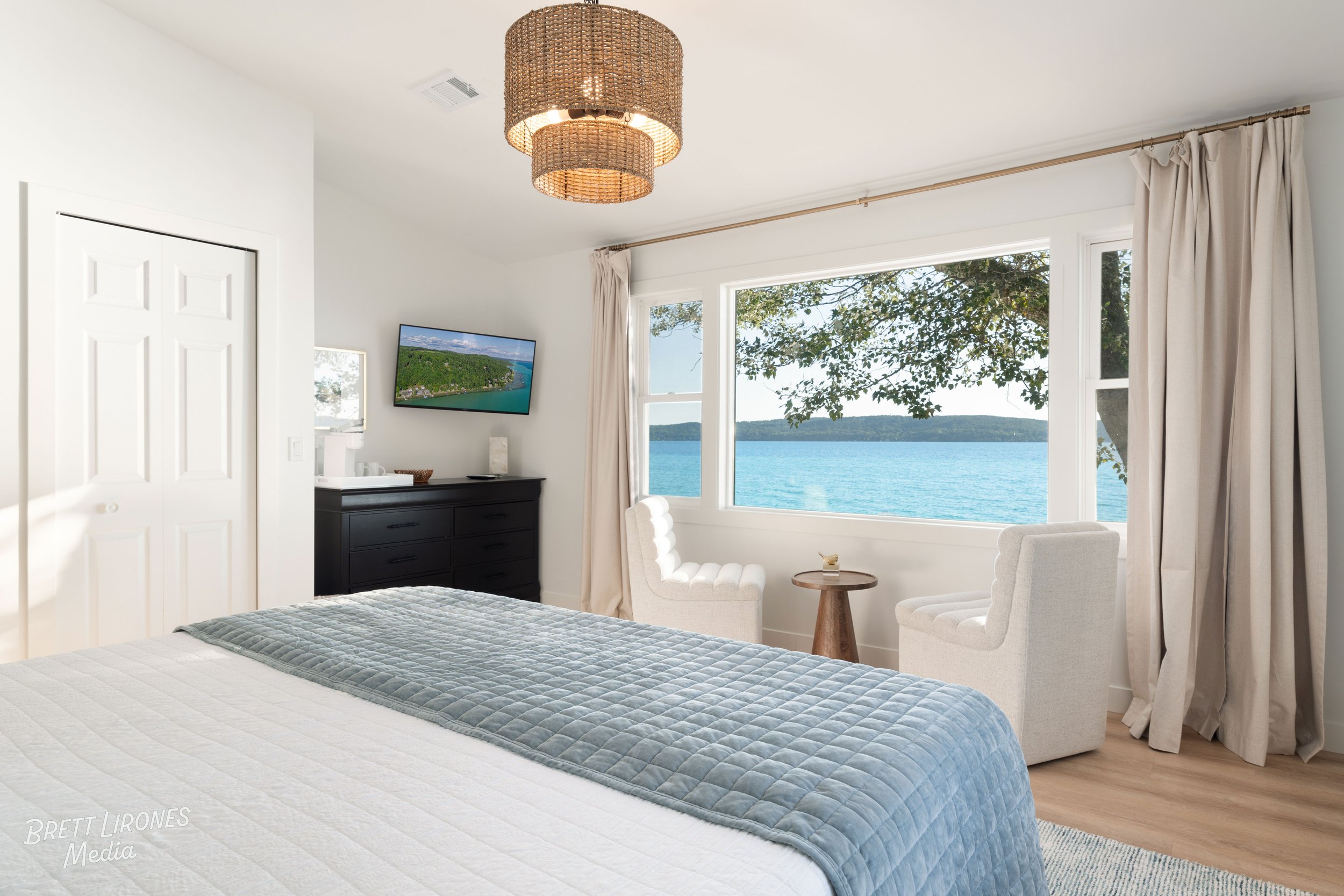 A bedroom with a large window overlooking a body of water and trees, two white armchairs and a small round wooden table in front of the window, a black dresser with a TV, and a bed with a white and gray quilt.