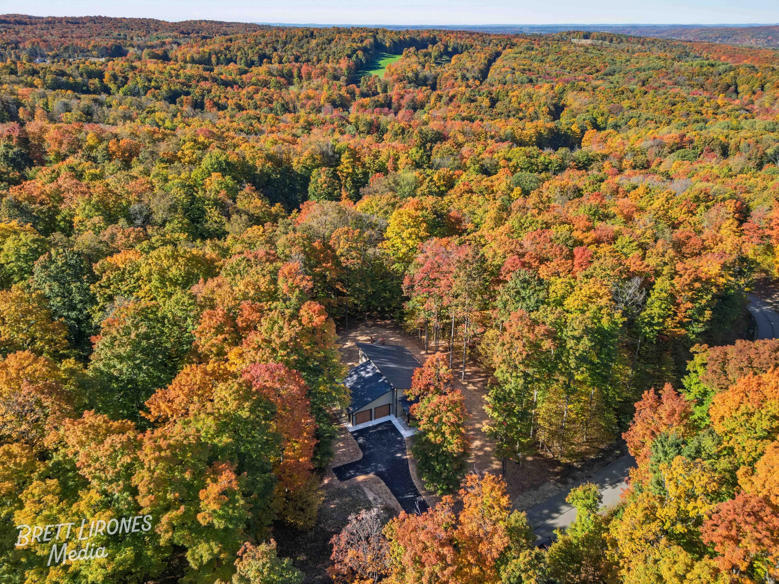 An aerial view of a forest with fall foliage in shades of red, orange, yellow, and green. A house with a dark roof and a driveway is visible among the trees. Rolling hills extend into the distance under a clear sky.