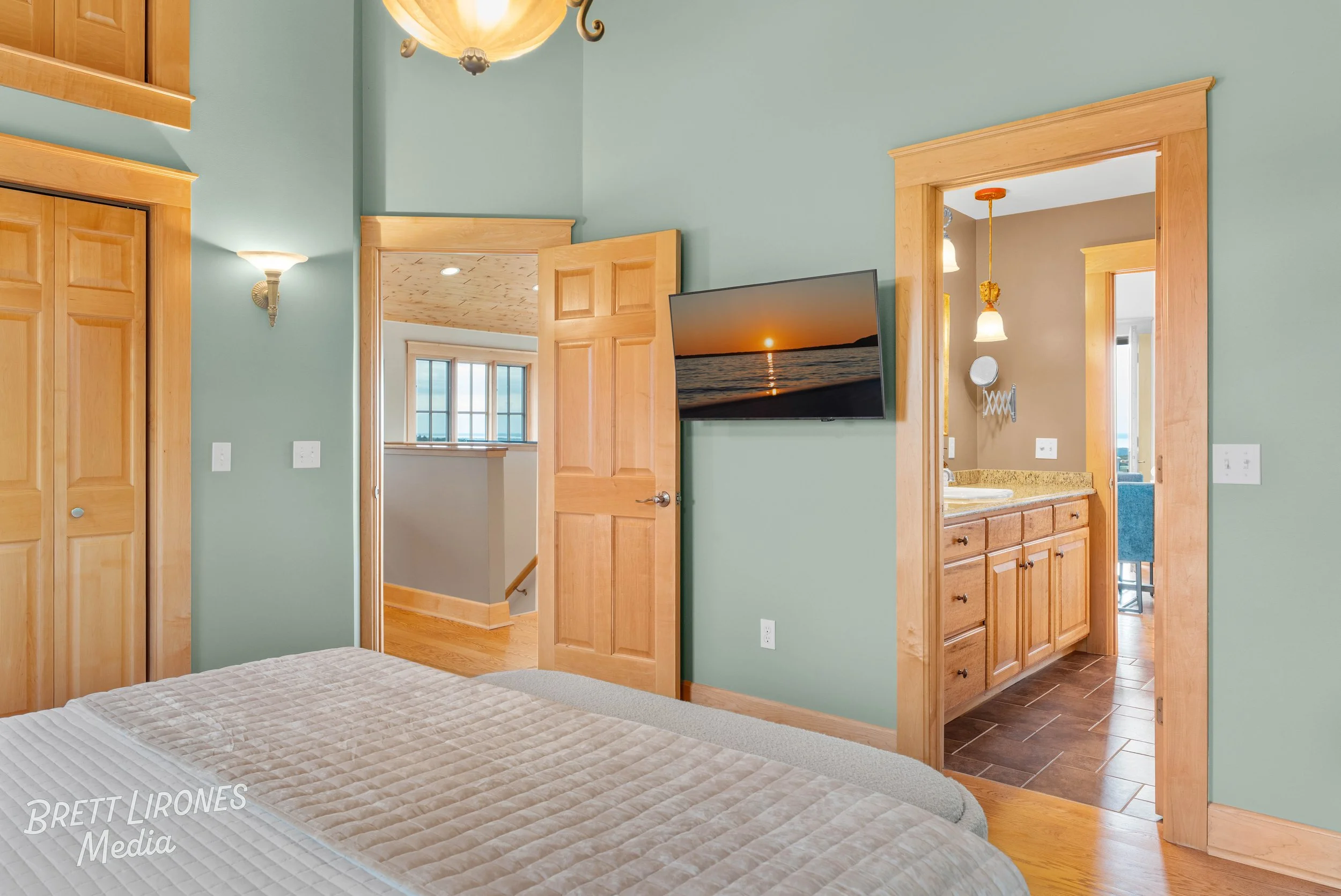 A bedroom with green painted walls, wooden door frames, a TV mounted on the wall showing a sunset over water, a bed with beige bedding, and an en-suite bathroom with wooden cabinetry and a mirror.