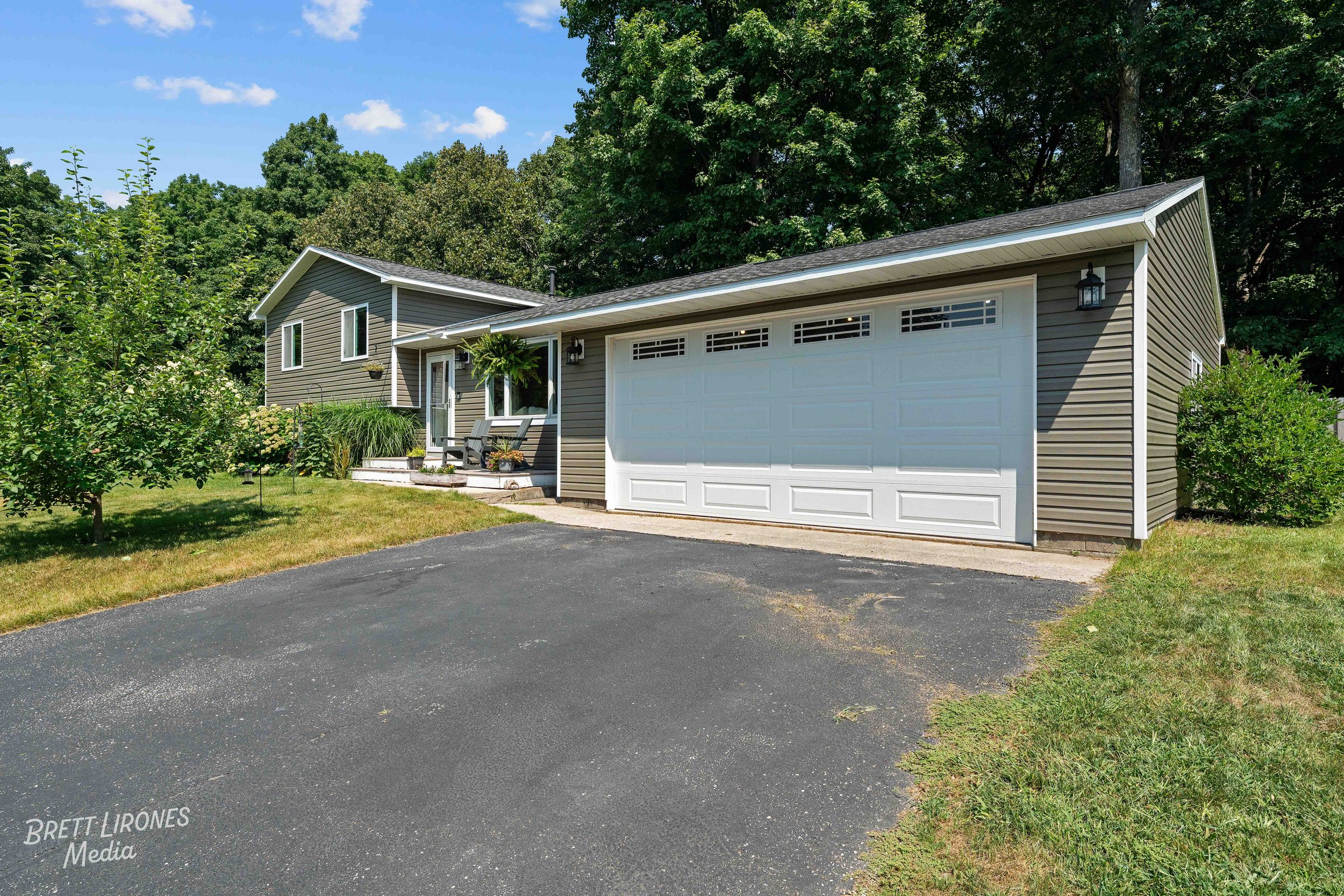 A house with gray siding, a white garage door, and a front porch with chairs, surrounded by greenery and trees, under a blue sky with some clouds.