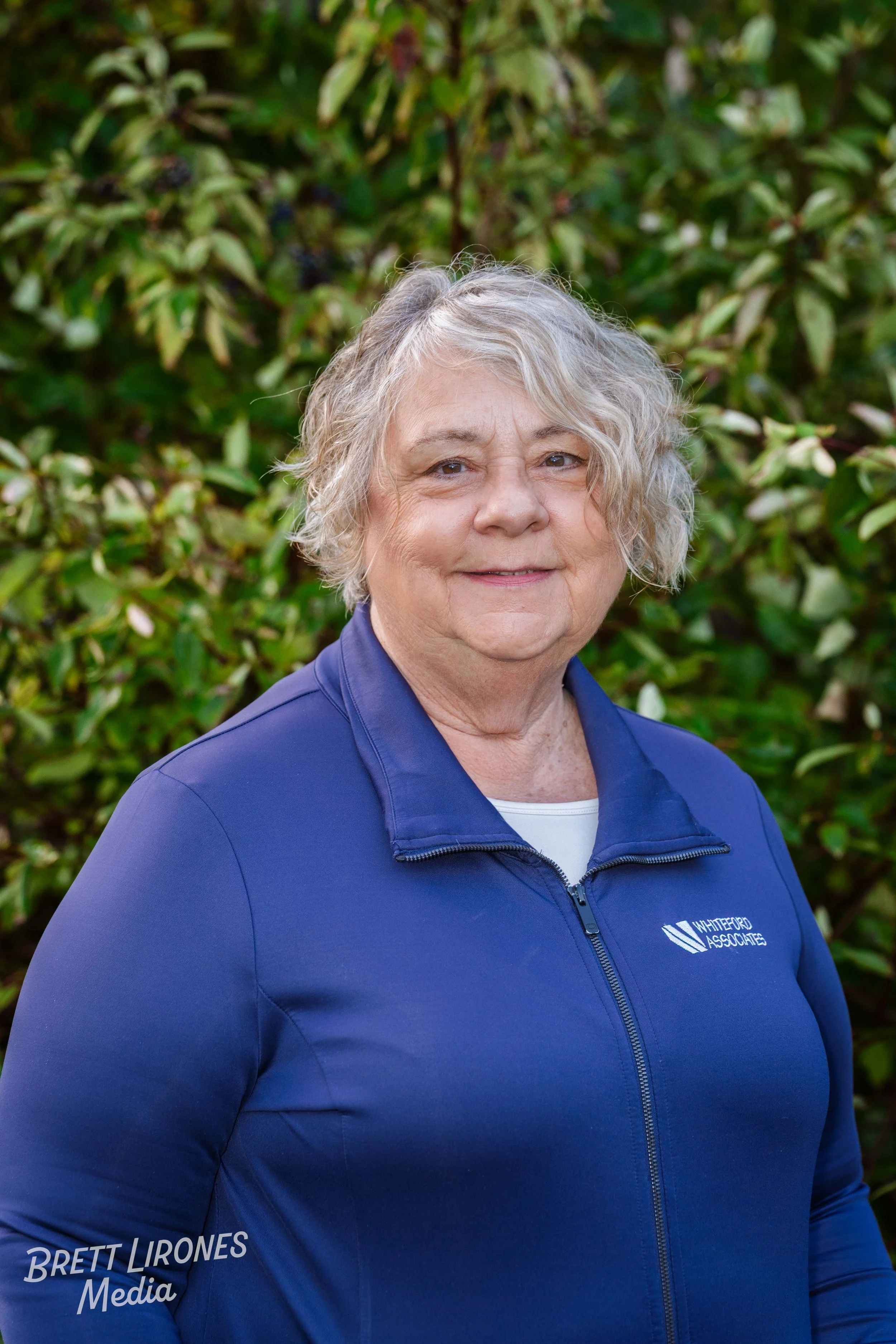 A smiling older woman with curly gray hair standing outdoors in front of green bushes, wearing a blue jacket with a logo that reads 'Whiteford Associates'.
