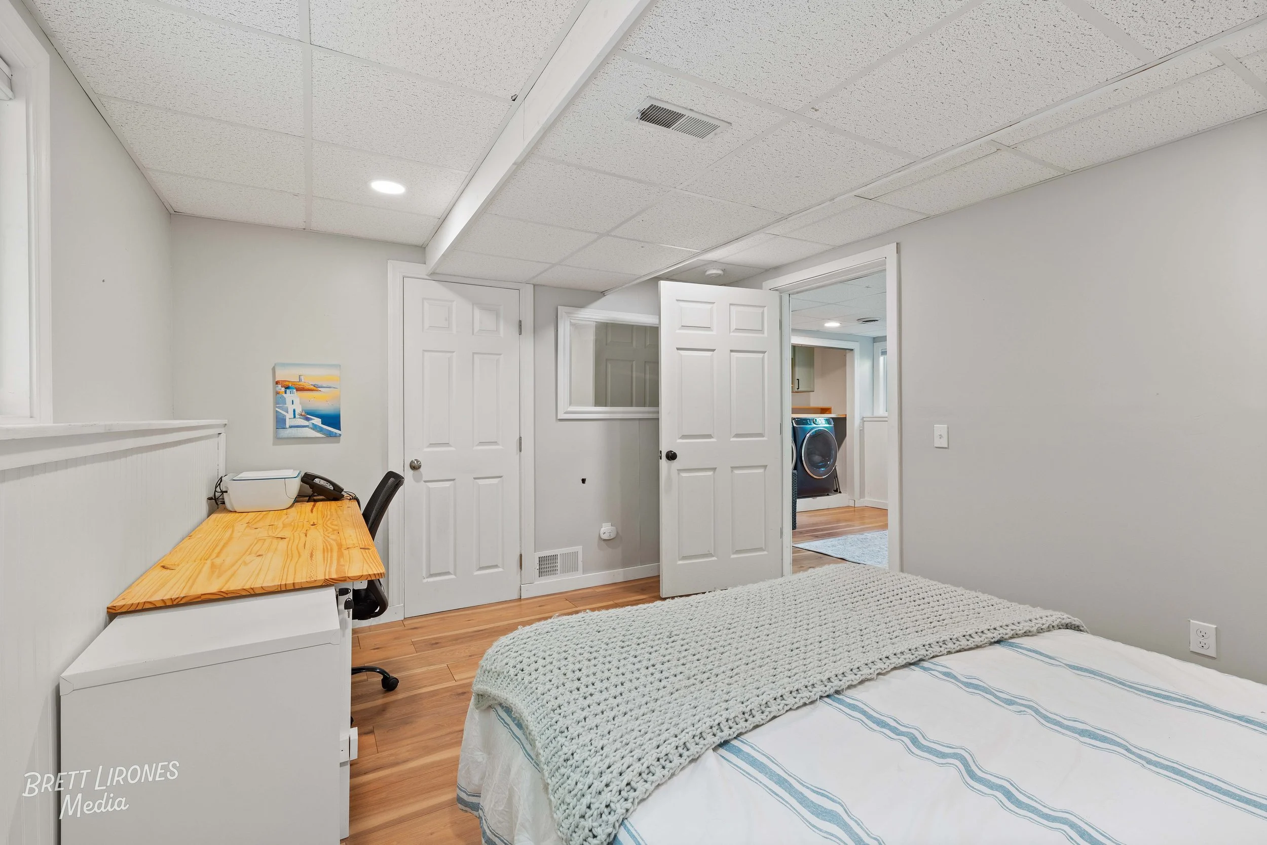 A bedroom with a bed, a wooden desk, and an office chair, with doors leading to a laundry area with a washing machine.