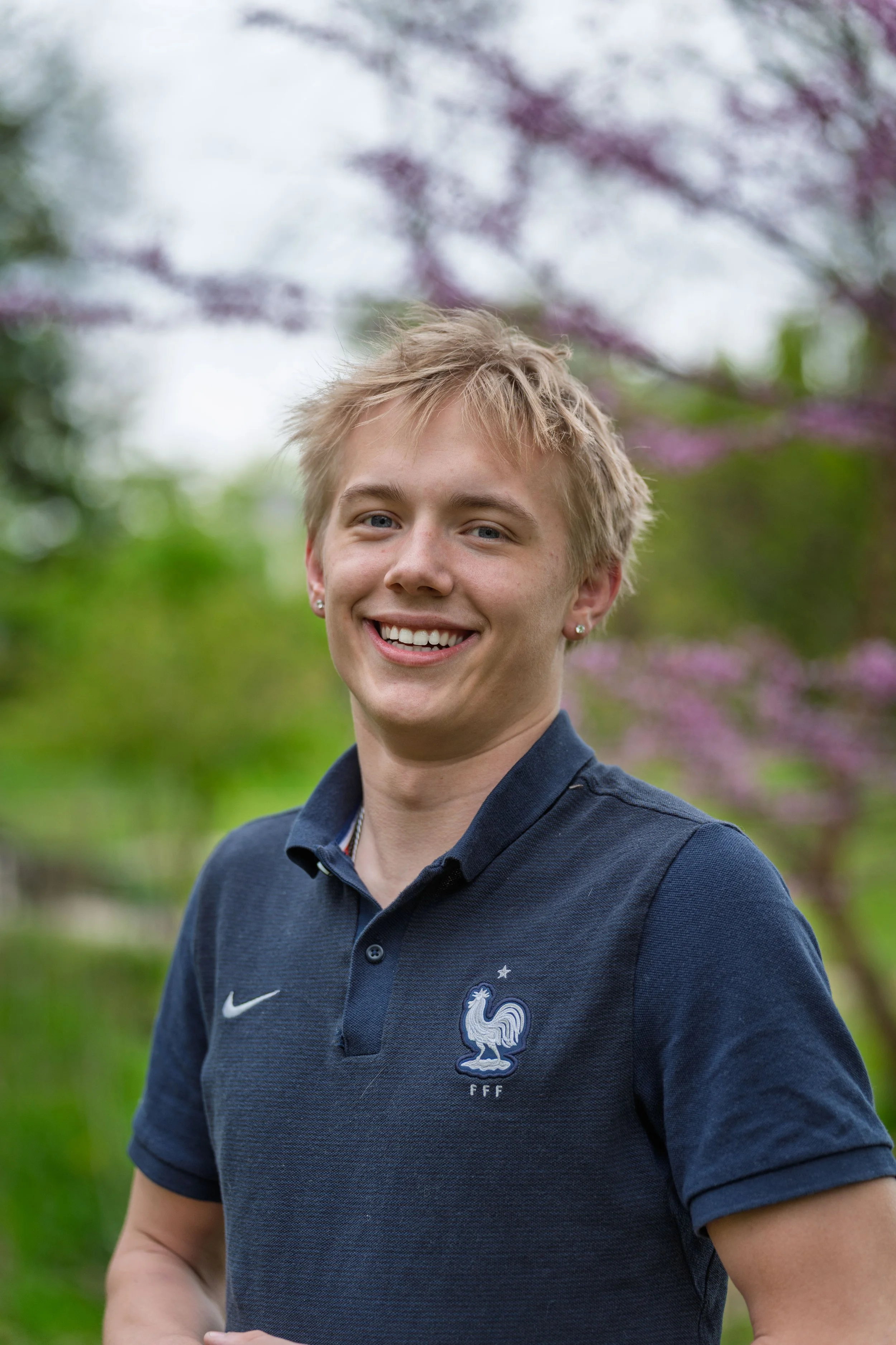 A young man smiling outdoors with trees and pink flowers in the background, wearing a navy blue polo shirt with an embroidered rooster logo.