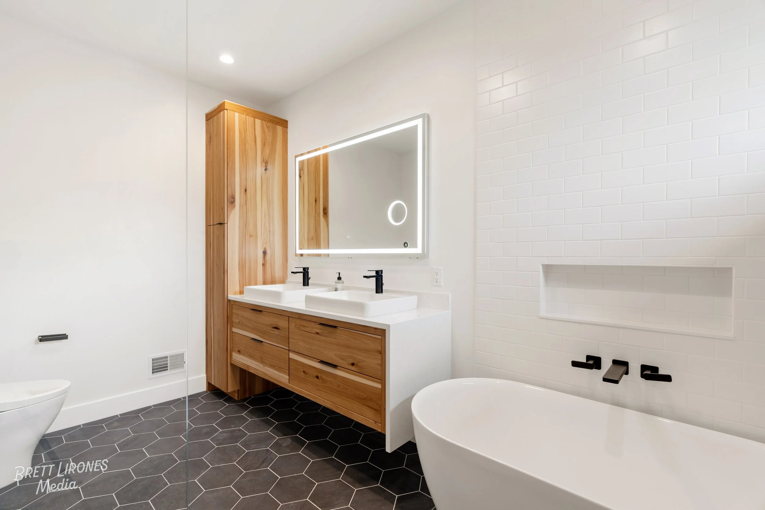 Modern bathroom with white walls, a black hexagon-tiled floor, wooden vanity with two sinks, large illuminated mirror, and a freestanding bathtub.