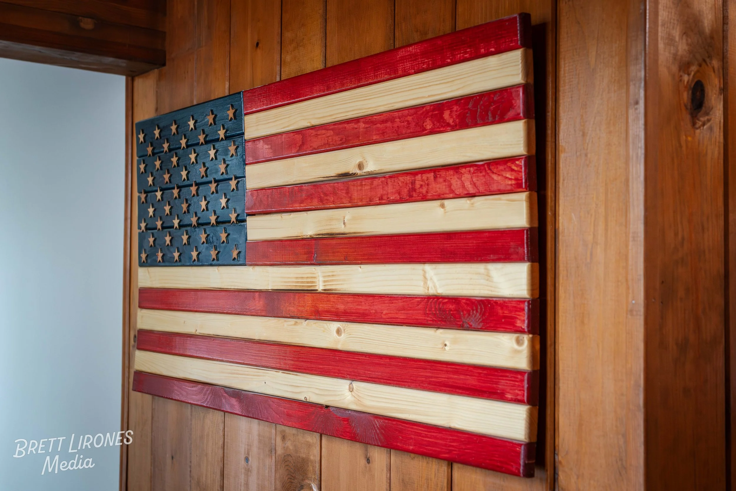 Wooden American flag decor with natural and red painted stripes and a blue canton with cut-out stars, placed on a wooden surface.
