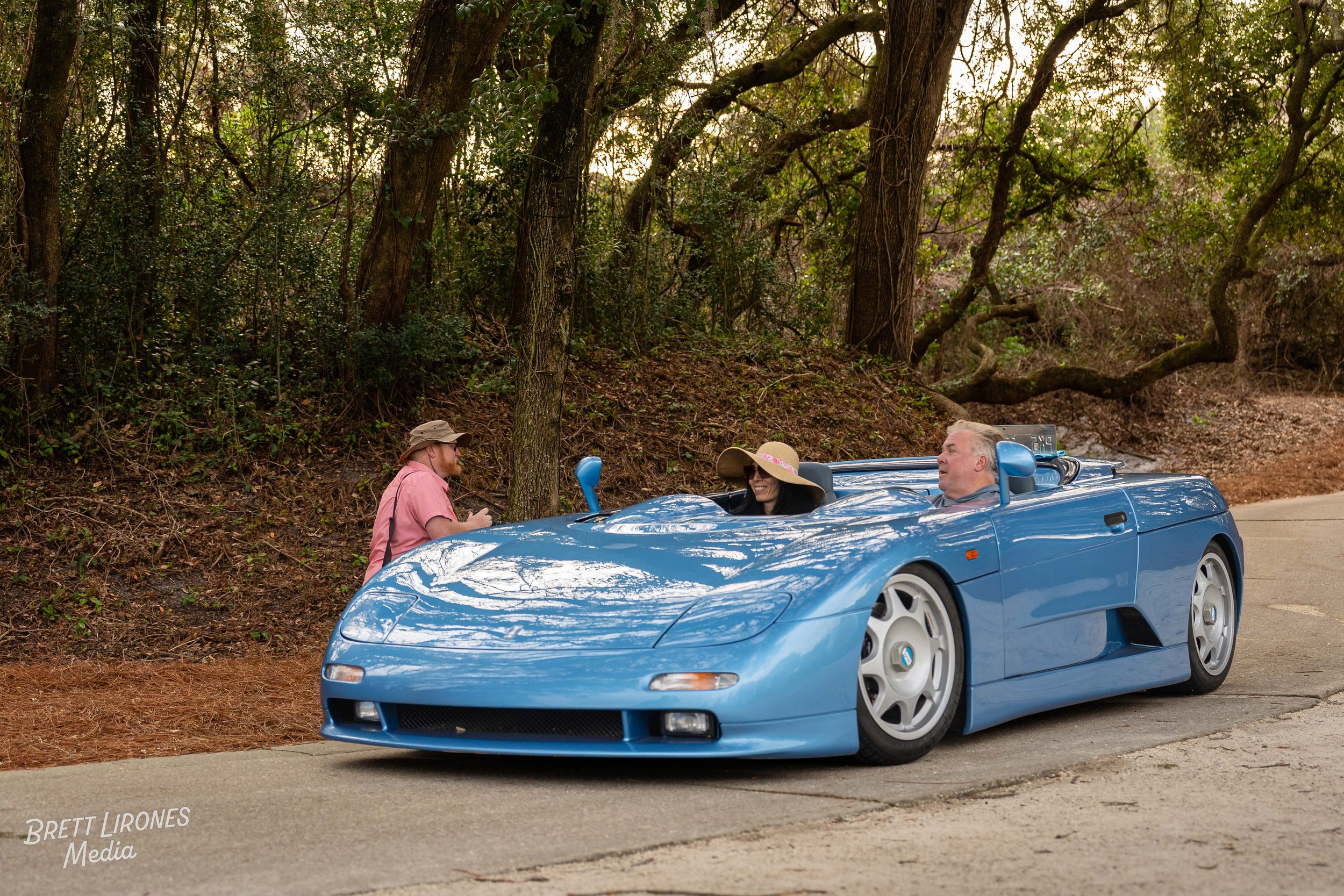 A blue sports car with two people inside, parked on the side of a road near a wooded area, with a person standing beside it talking to the occupants.