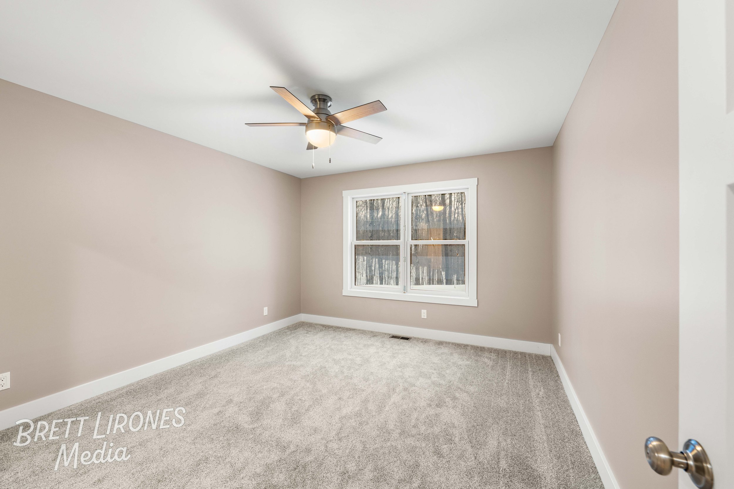 Empty room with beige walls, white trim, a window showing trees outside, a ceiling fan, and light gray carpet.