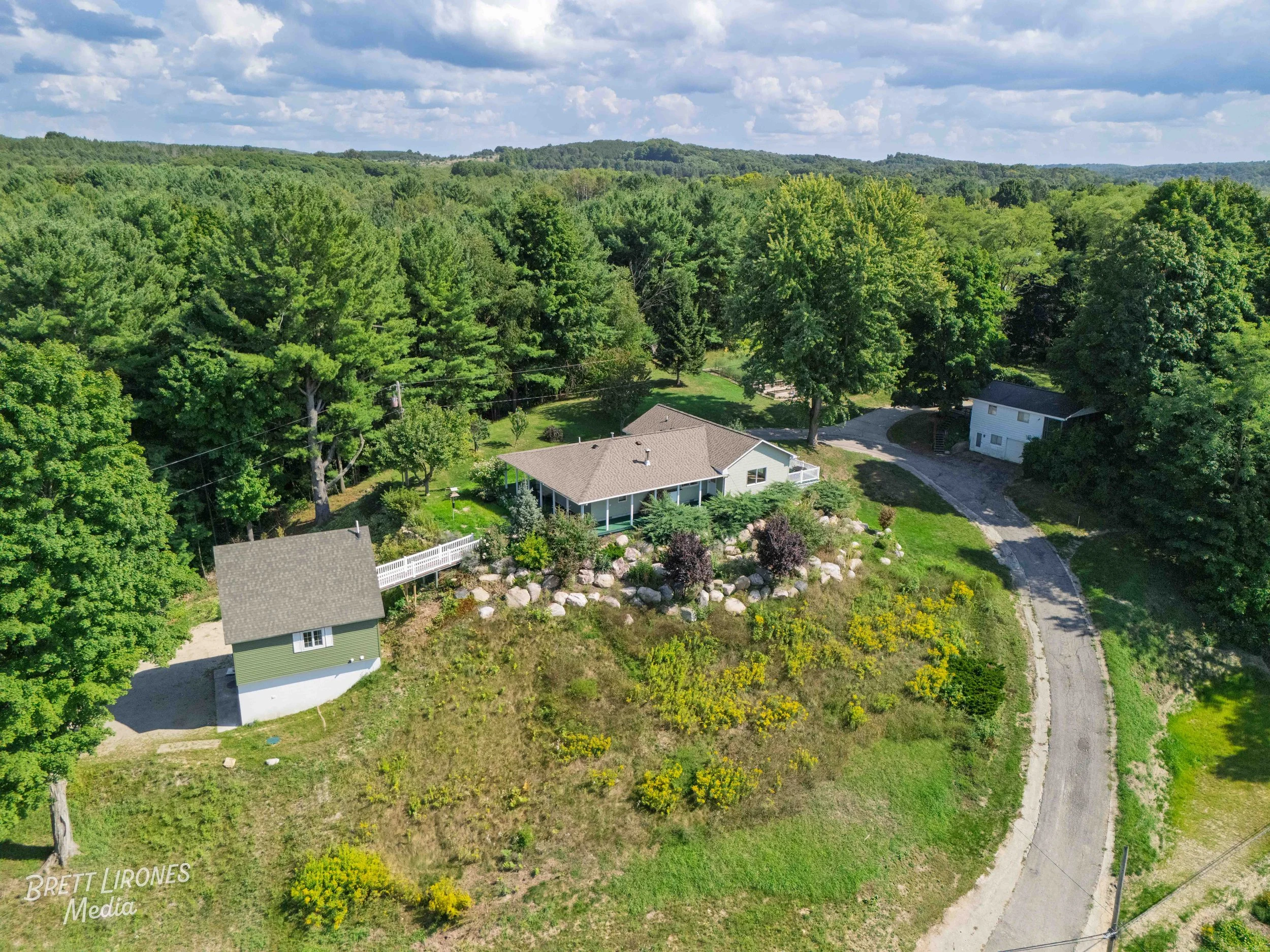 An aerial view of a house surrounded by lush green trees, with a curved driveway leading to the house and a small detached building nearby, set in a forested area under a partly cloudy sky.