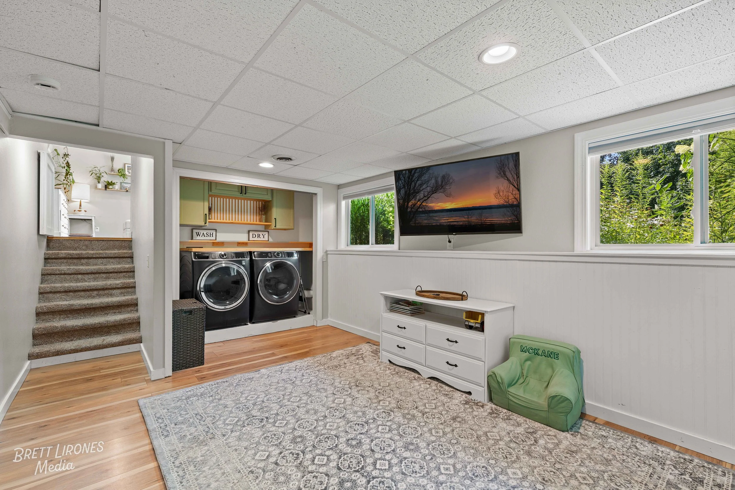 Laundry room with front-loading washer and dryer, white walls, windows with greenery outside, a wall-mounted TV, a white dresser, and a green toy chair.
