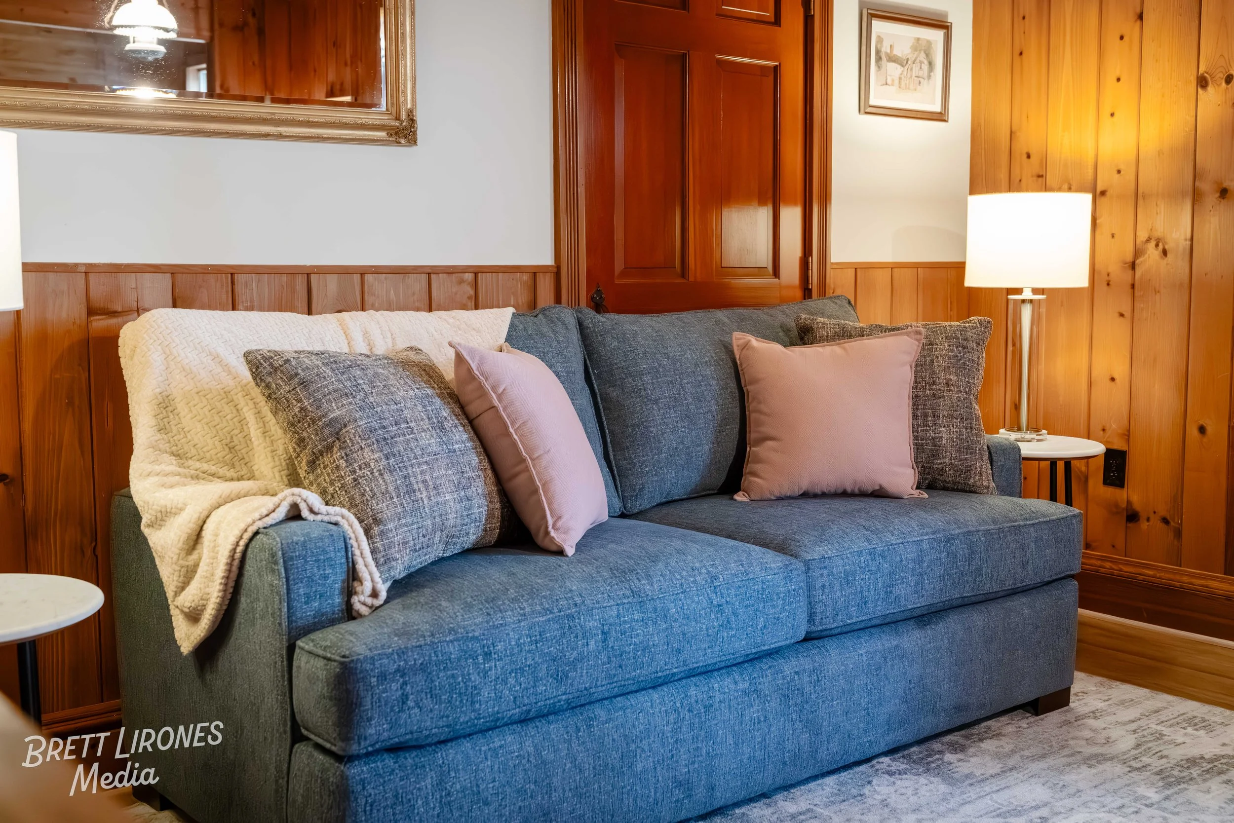 A cozy living room corner with a dark gray fabric sofa, pink and gray throw pillows, a cream knitted blanket, a wooden-paneled wall, and a side table with a white lamp.