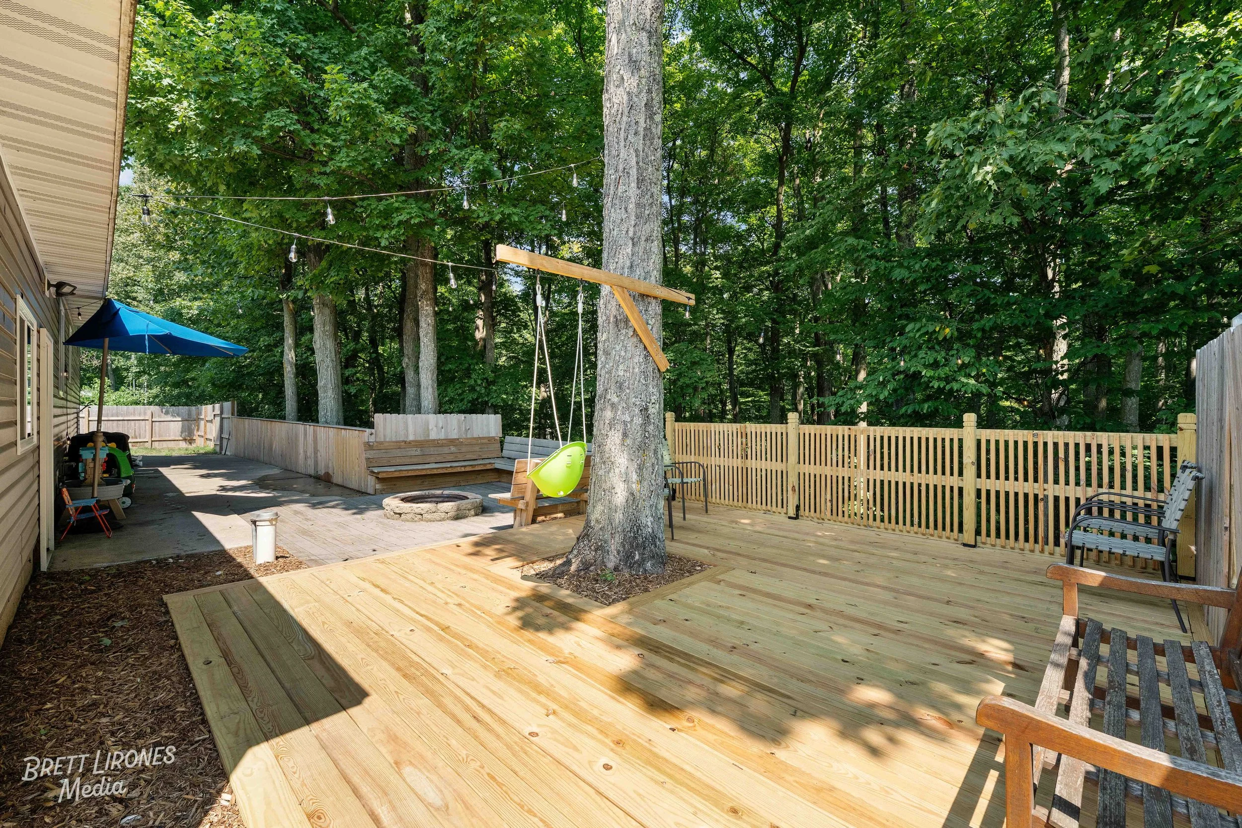 Backyard with wooden deck, swings with a green seat, benches, a fire pit, a blue umbrella, and string lights, surrounded by trees and wooden fencing.
