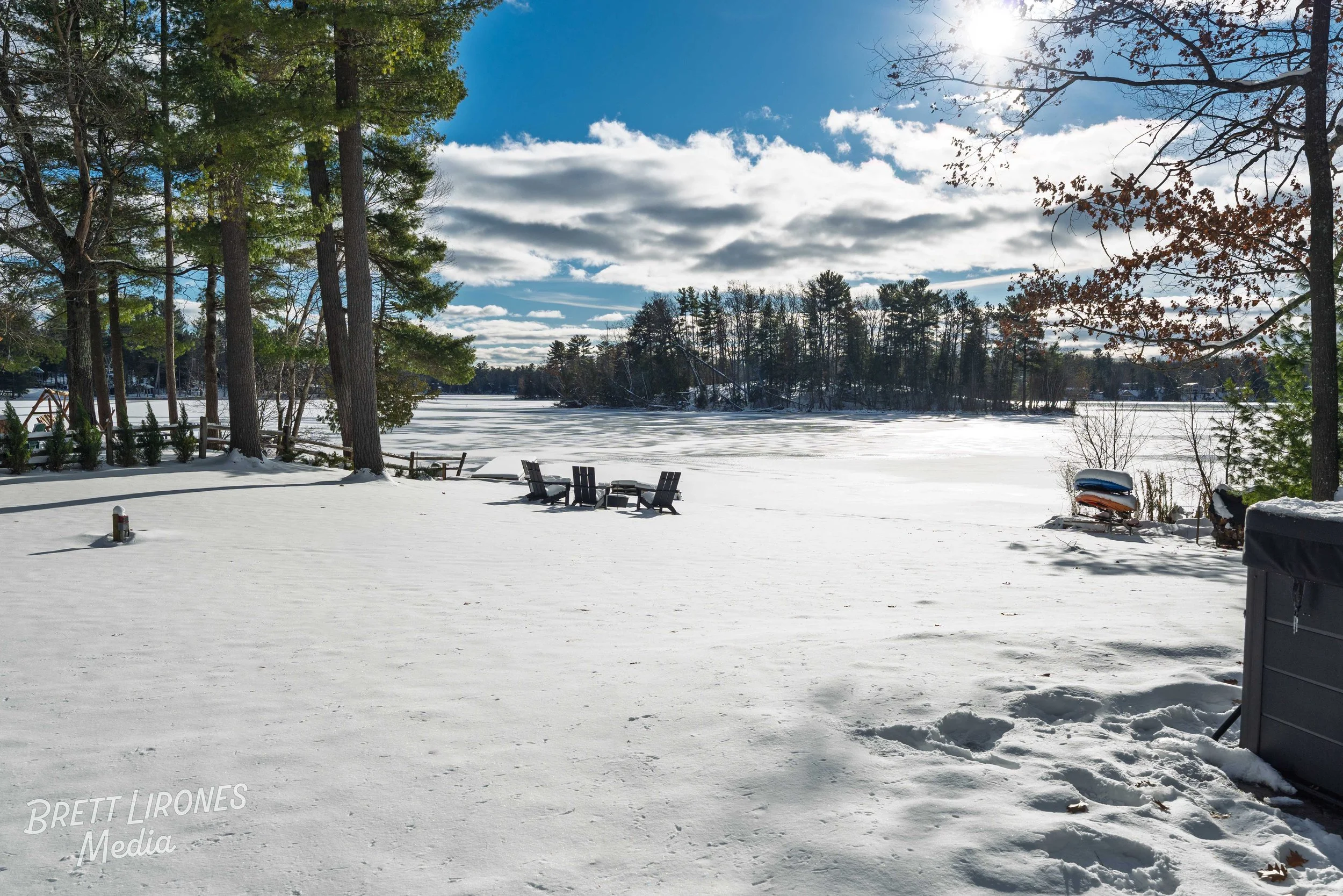 Snow-covered backyard with wooden chairs and a table, surrounded by trees and a frozen lake in the background, under a partly cloudy sky with the sun shining.