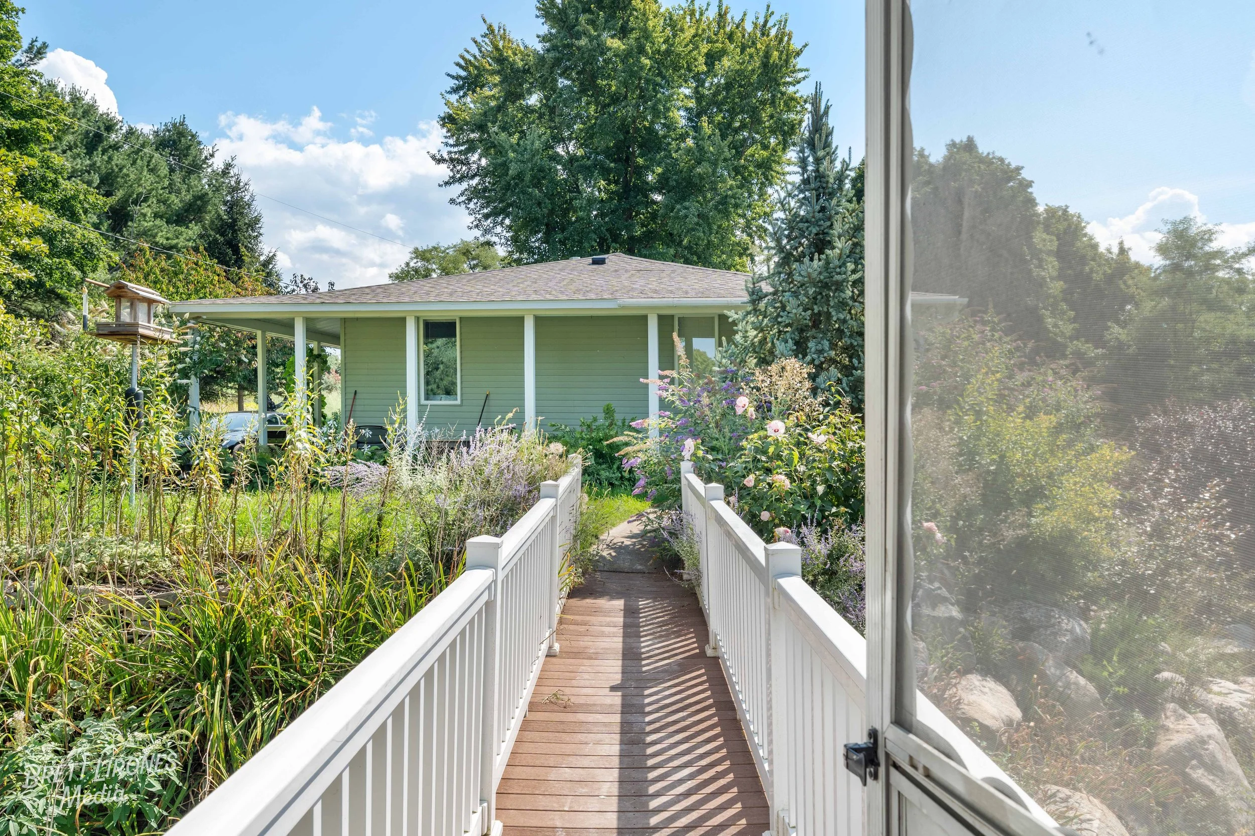 View from a screened porch leading to a backyard garden with various plants, a white house with a porch, surrounded by lush trees and a partly cloudy sky.
