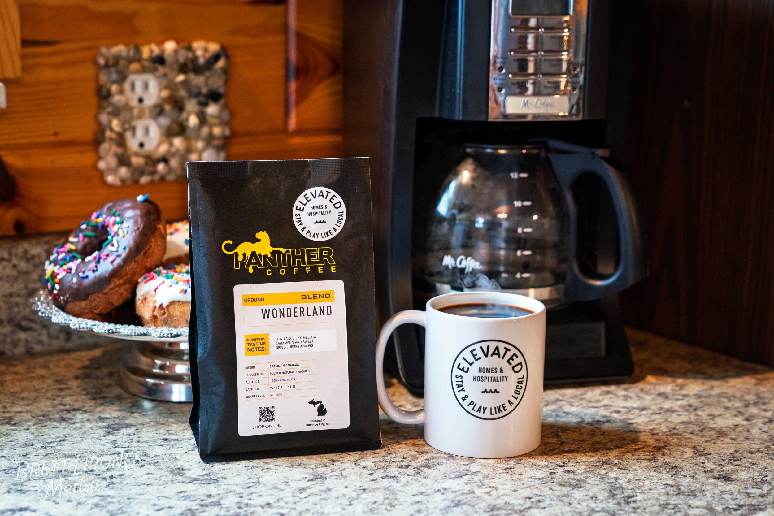 A cup of coffee, a bag of Panther Coffee Wonderland blend, and a plate of assorted donuts on a counter with a coffee maker in the background.