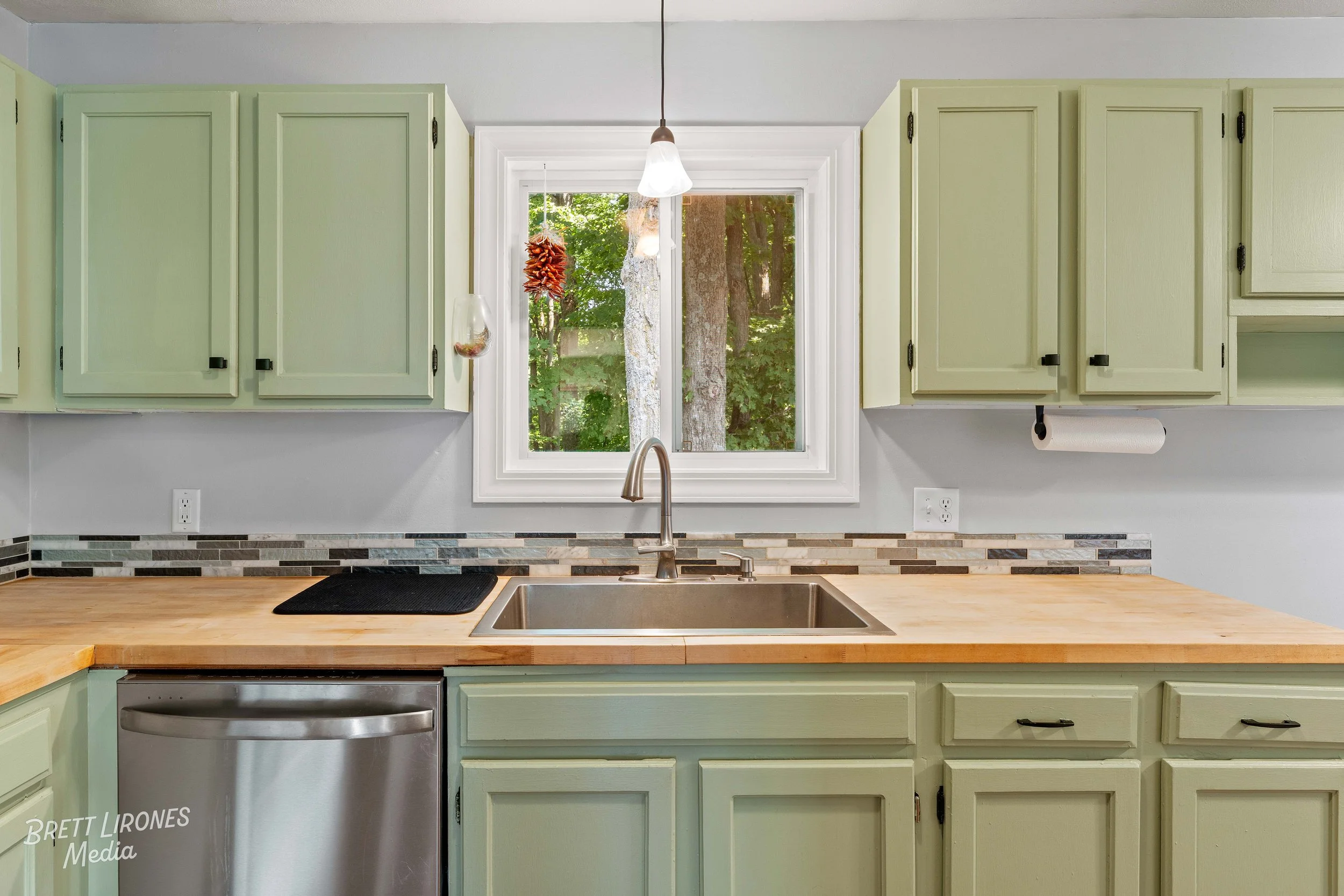 Kitchen with light green cabinets, a wooden countertop, a stainless steel sink, and a window showing trees outside.