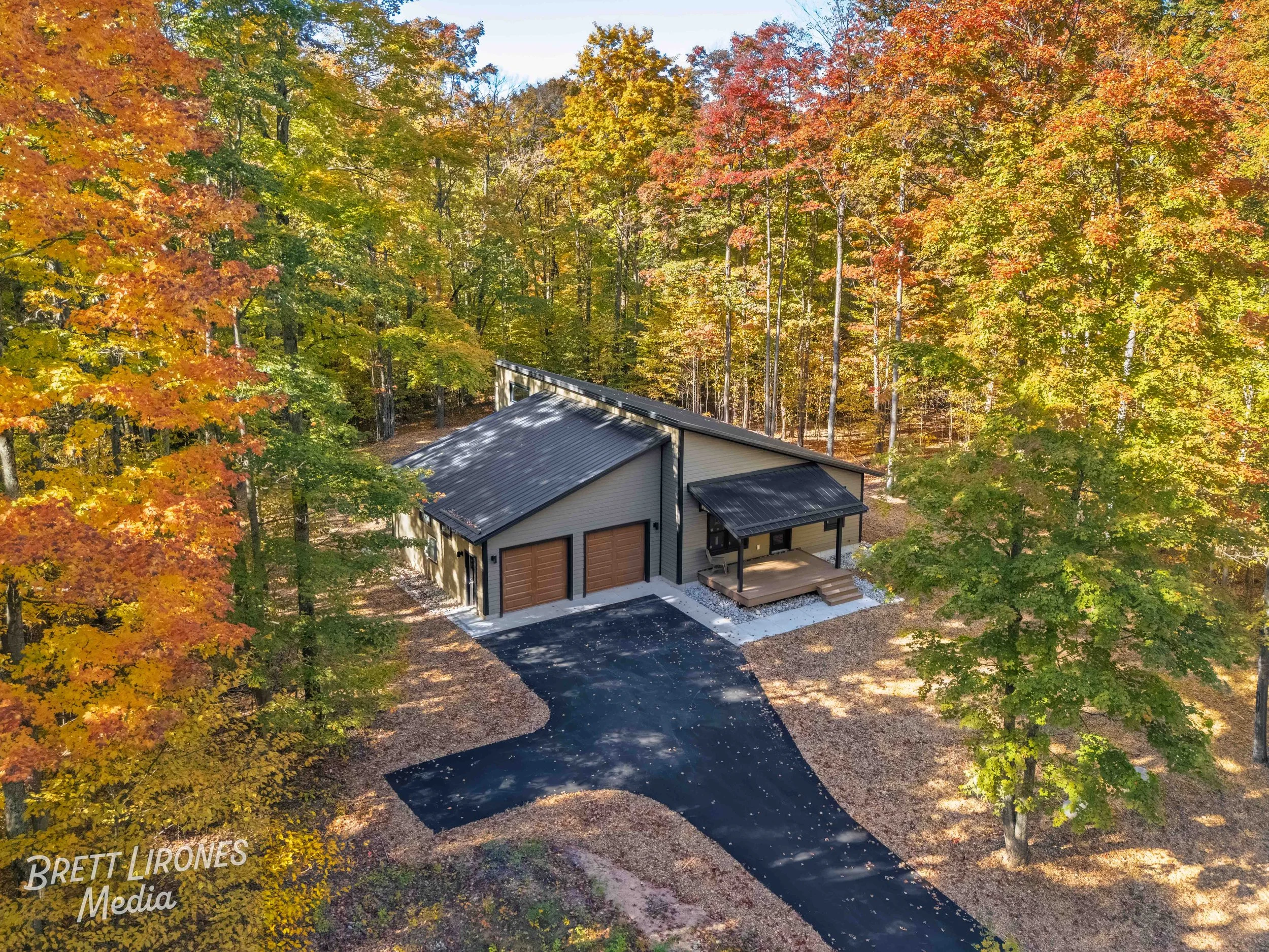 A modern, small house located in a wooded area with fall foliage, featuring a black metal roof, beige siding, a two-car garage, a small porch, and a paved driveway.