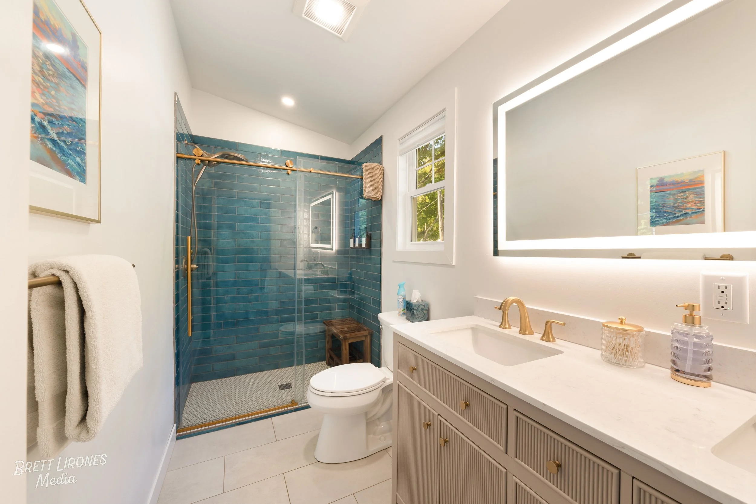 Modern bathroom with a shower area with teal tiles, a white toilet, a vanity with a marble countertop, a large illuminated mirror, and a window showing greenery outside.