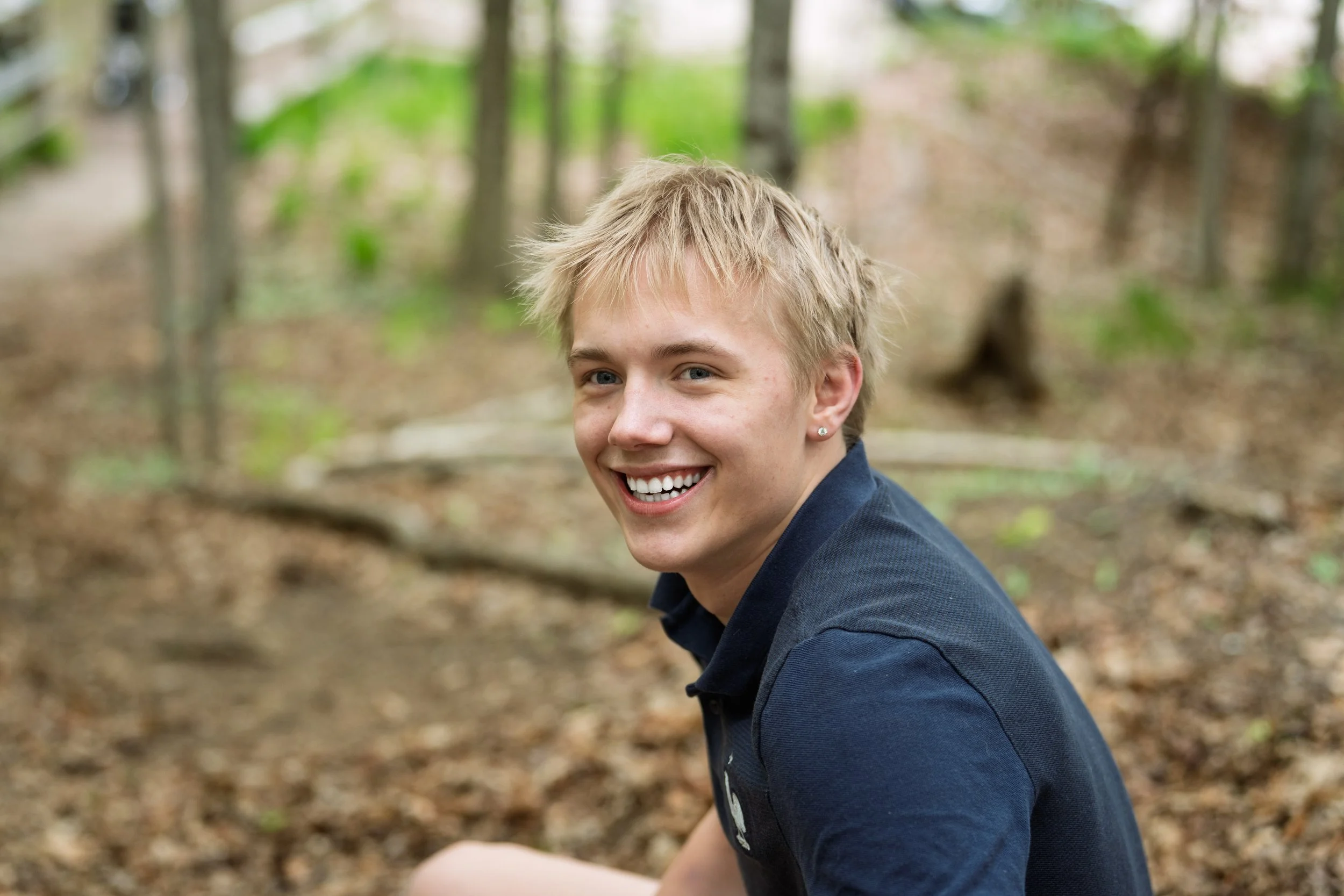 A young man with blond hair, smiling, sitting in a forested area.