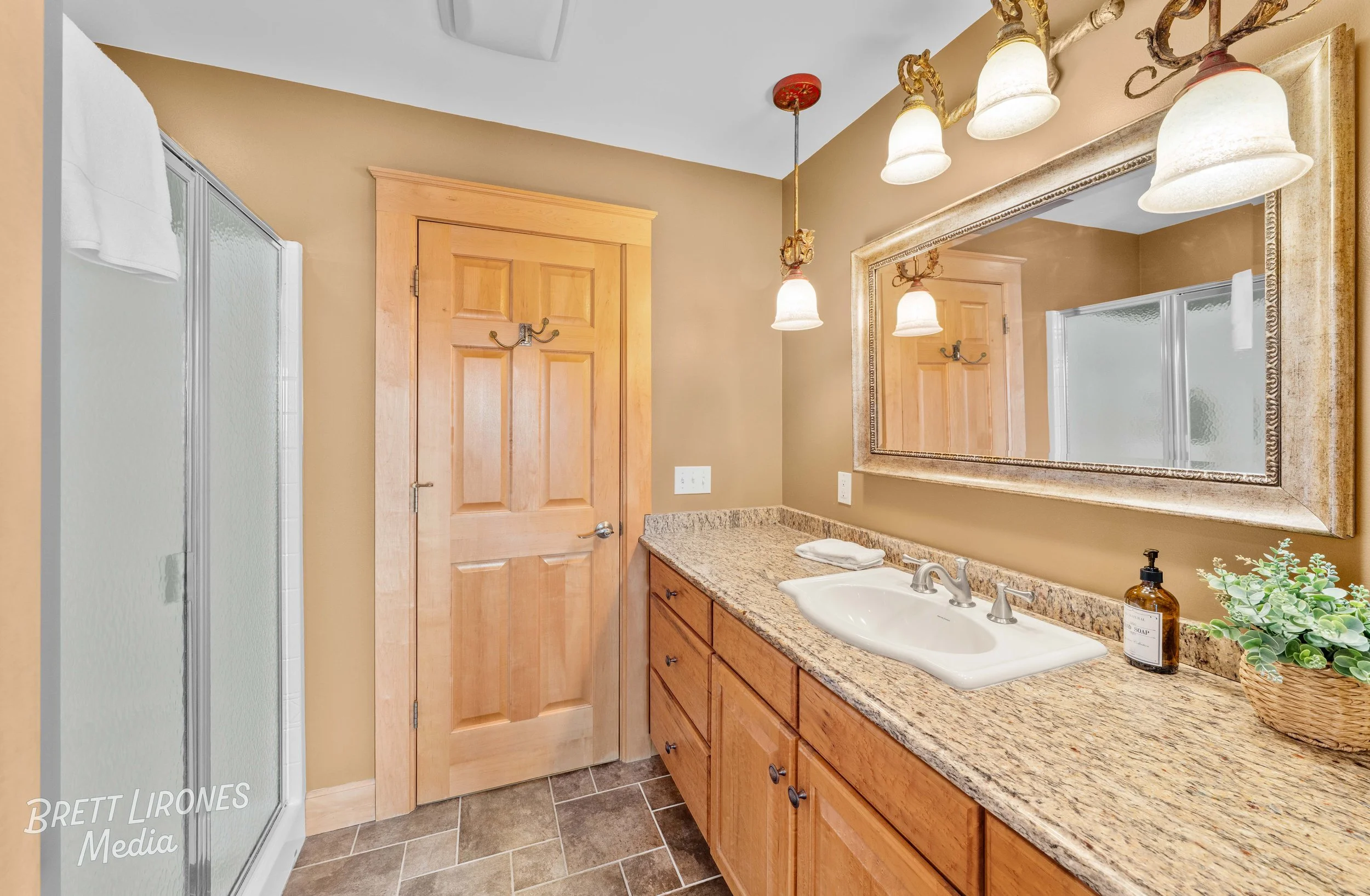 Bathroom with a granite countertop vanity, a large mirror, wall-mounted light fixtures, a wooden door, a shower with frosted glass, beige walls, and brown tile flooring.