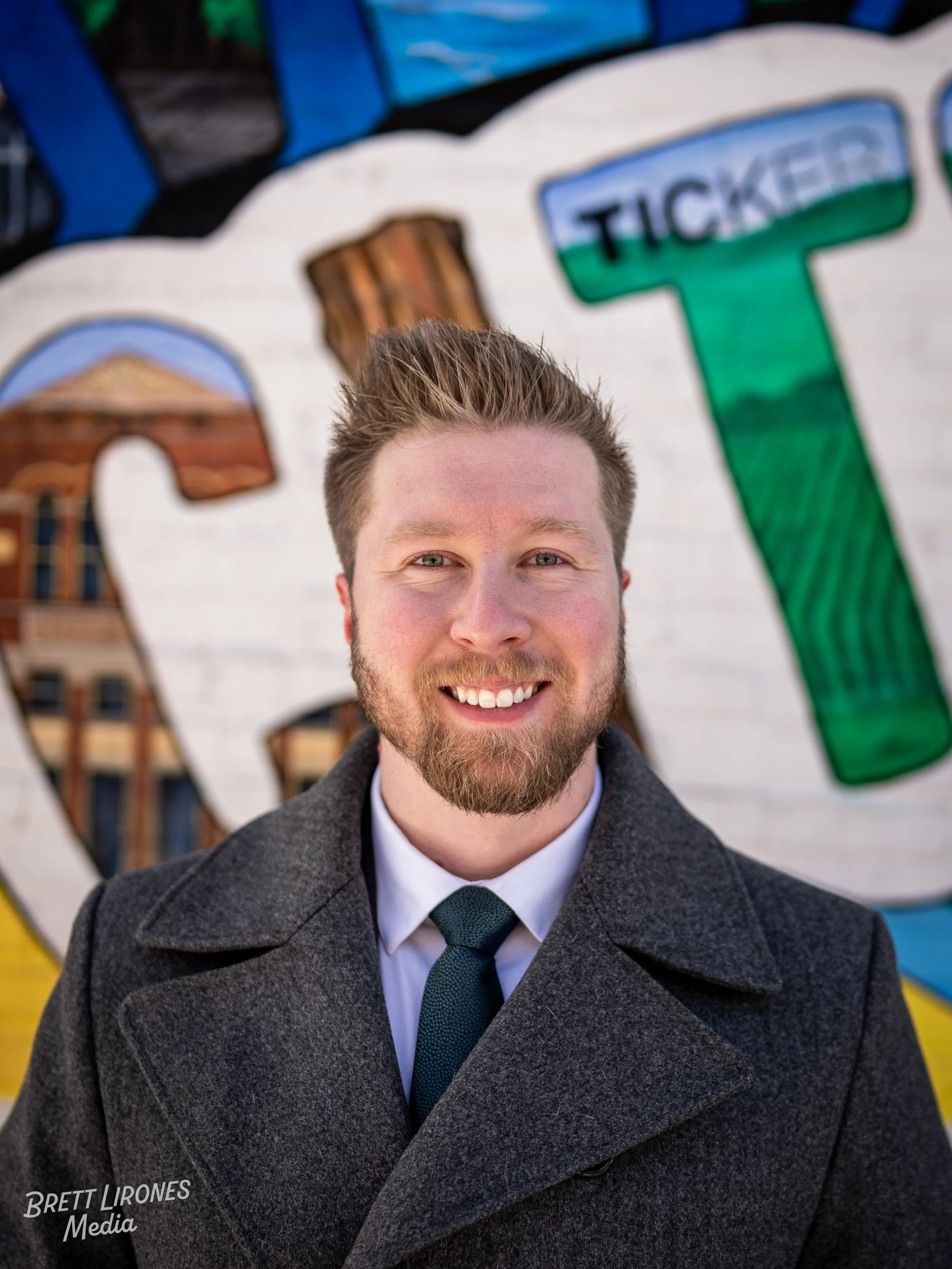 A smiling young man with a beard, wearing a gray coat, white shirt, and dark tie, standing outdoors in front of a colorful mural with street art and lettering.