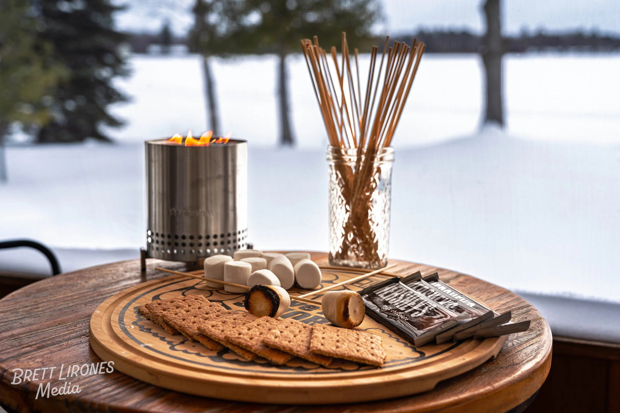 A wooden table with a round wooden serving tray holding graham crackers, marshmallows, chocolate bars, and roasting sticks. There is a metal candle warmer with a flame, and a glass jar filled with more roasting sticks. The background shows a snowy ou
