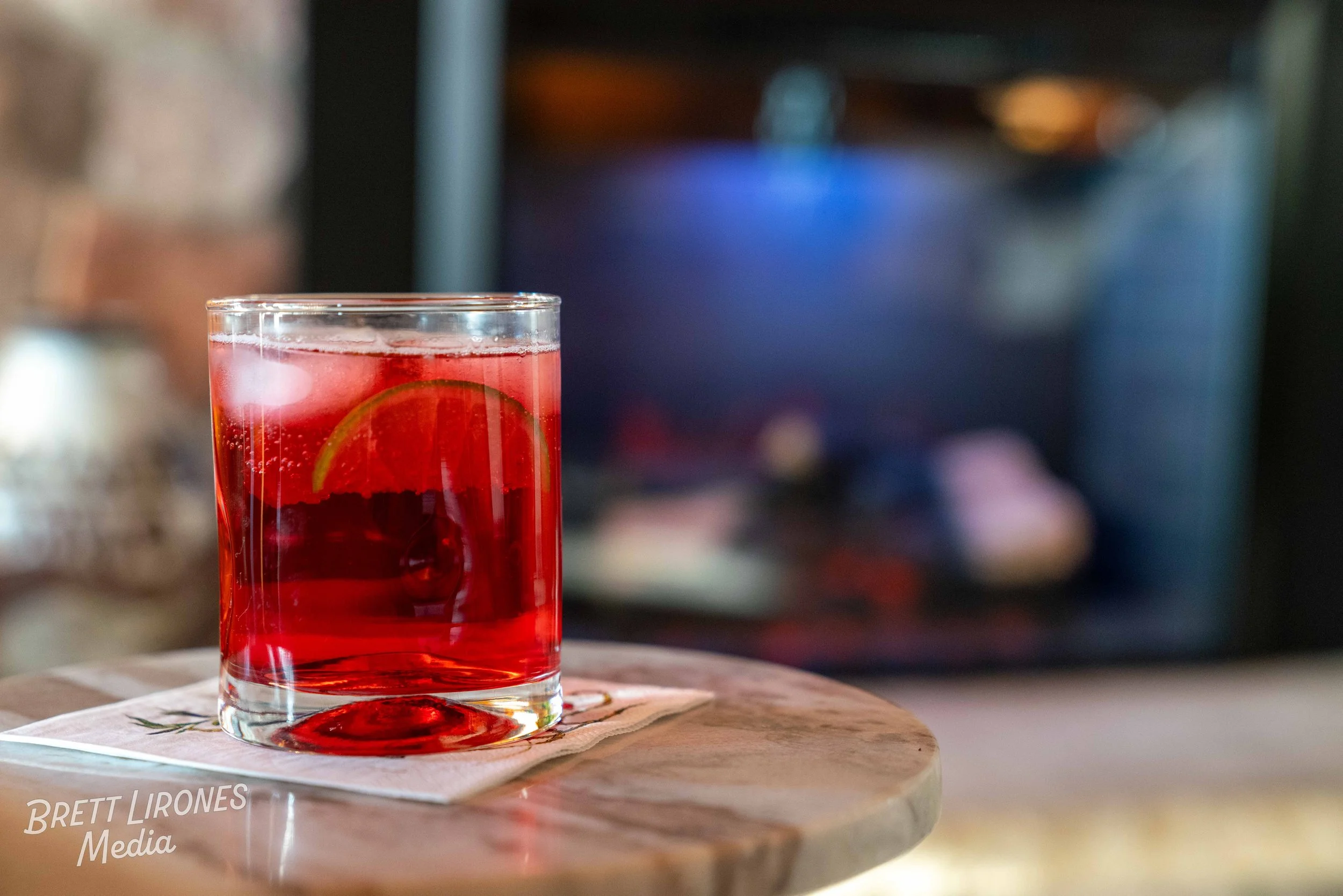 A glass of red beverage with lime slice and bubbles on a wooden table, blurred background with a fireplace