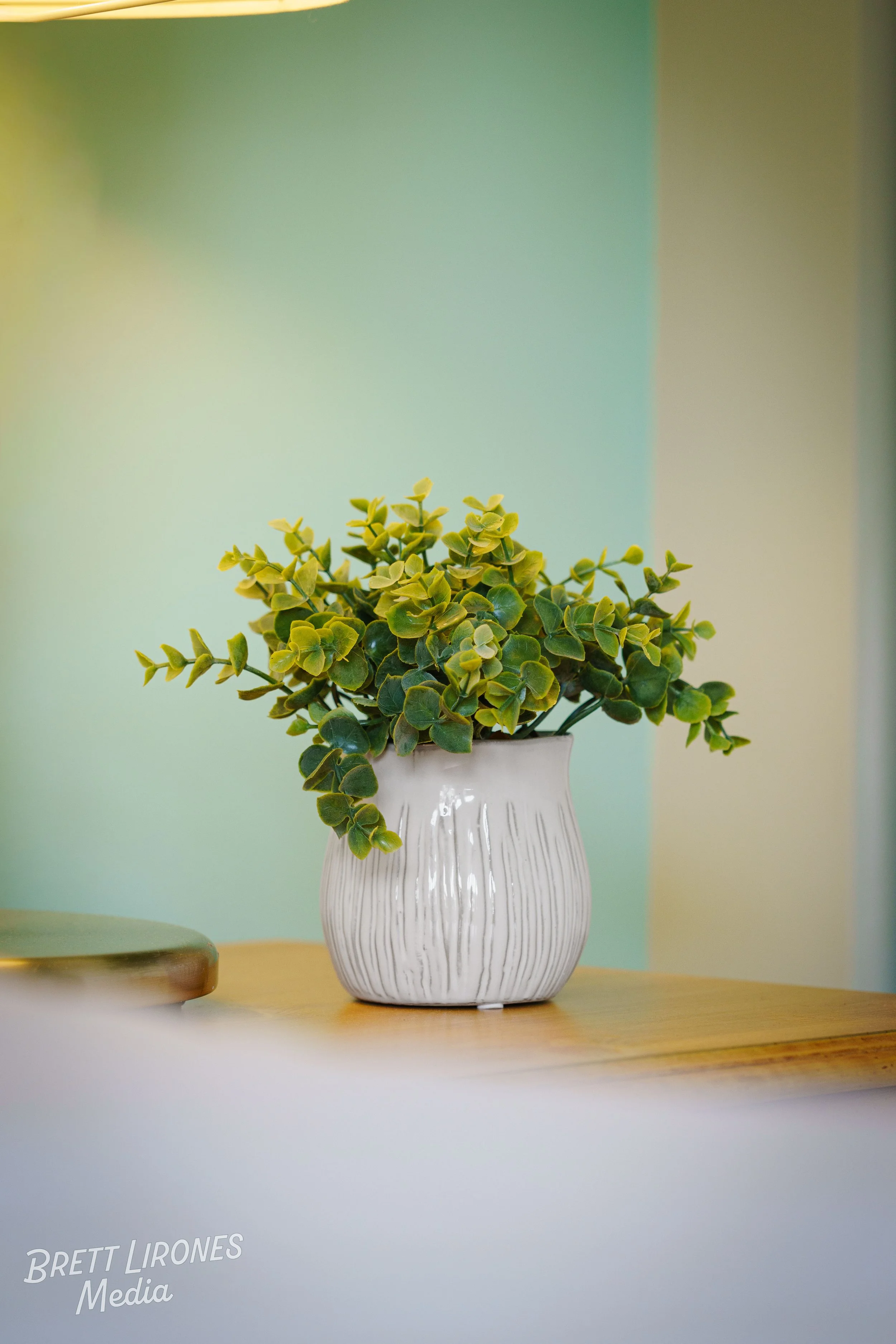 A white ceramic vase with vertical ridges, filled with a bouquet of green leaves and small yellow flowers, placed on a wooden surface against a blurred multicolored background.